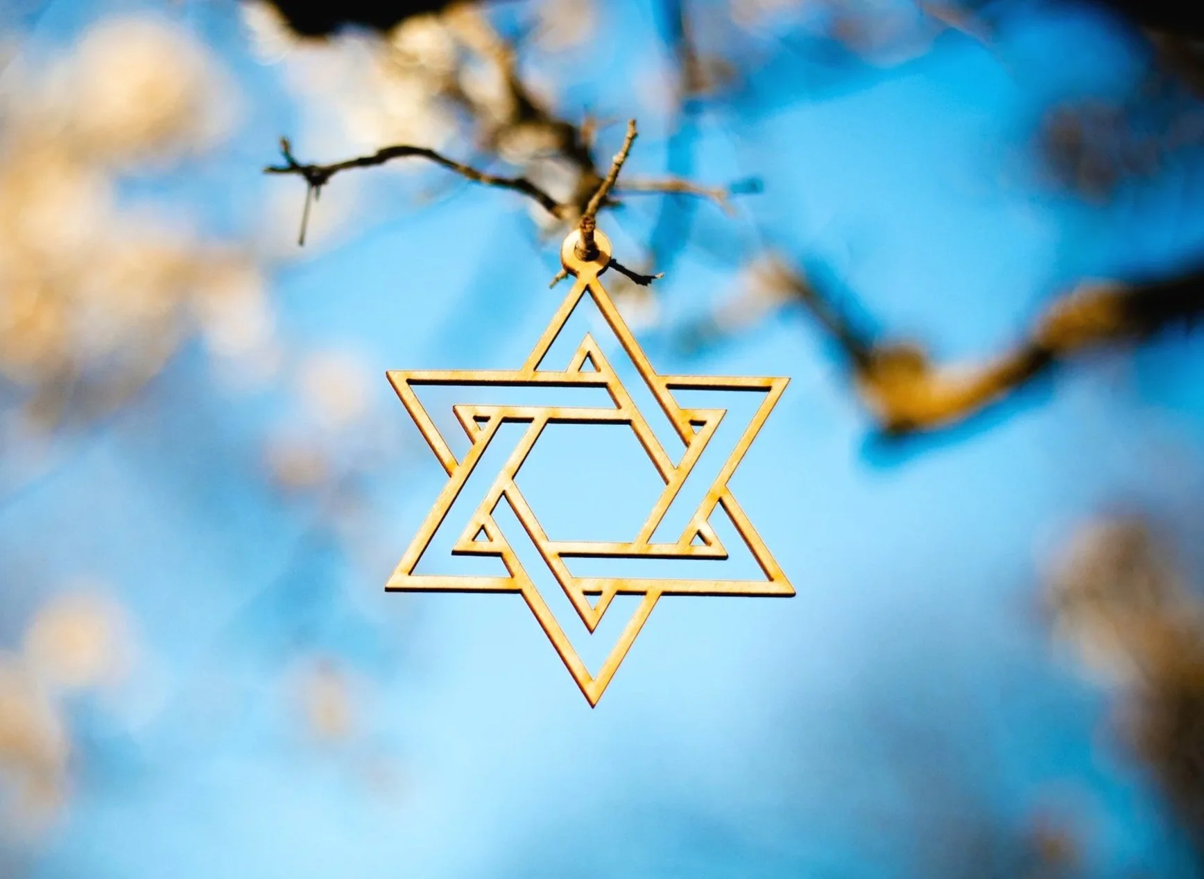 A wooden Star of David ornament hanging from a tree branch against a blue sky with some blurred clouds.