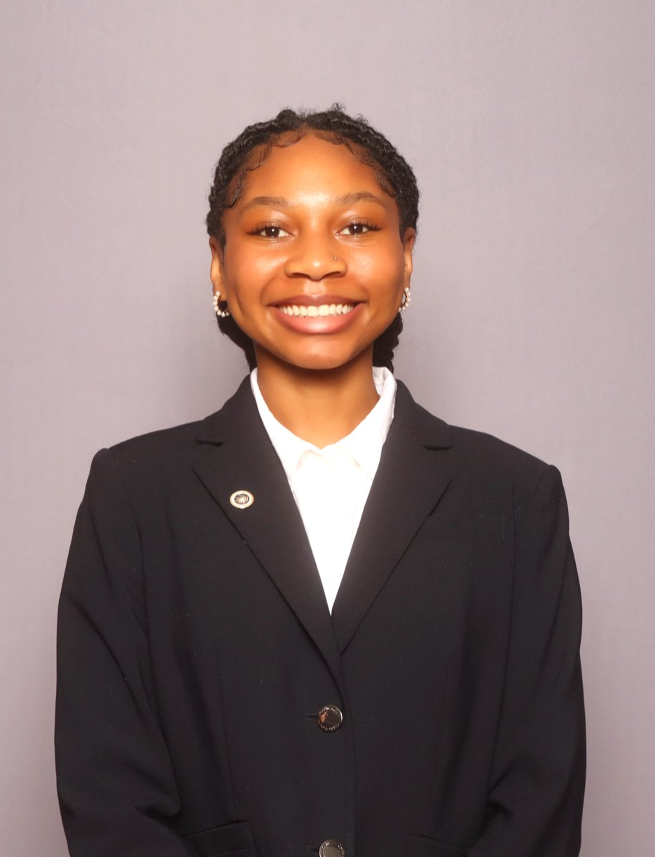 A young woman with short curly hair, wearing a black blazer and a white shirt, smiling in front of a gray background.