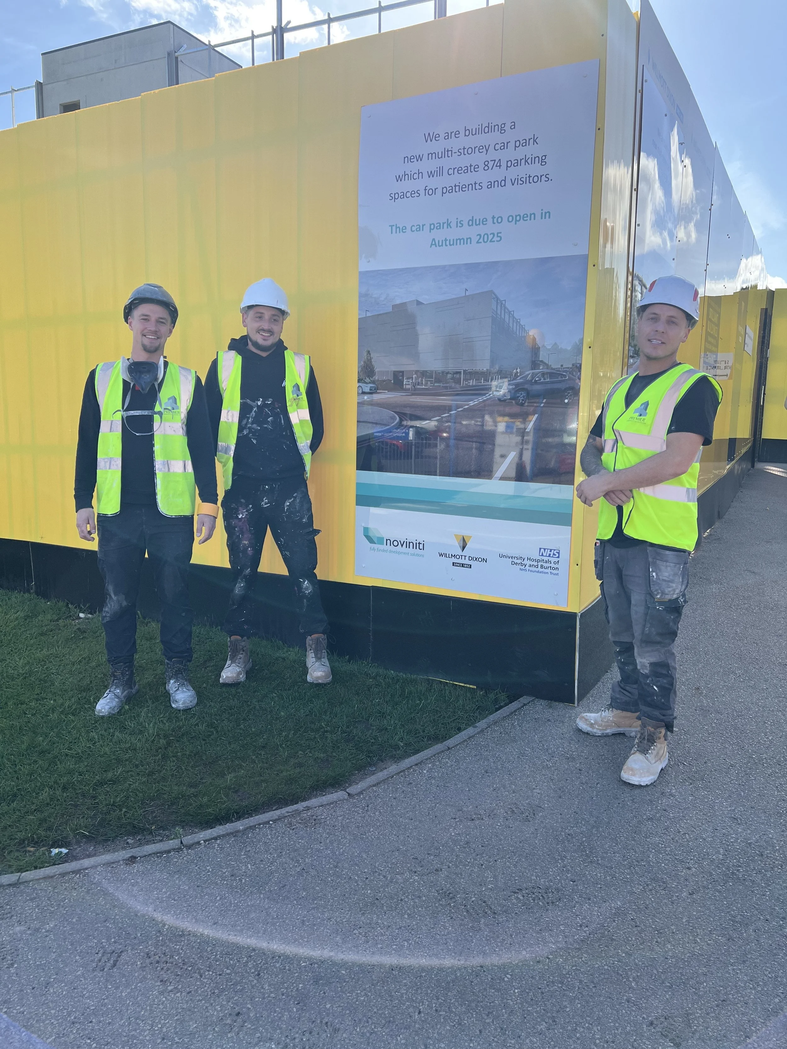 Three construction workers wearing high-visibility safety vests and helmets, standing outside near a construction site with a large yellow barrier and a sign about a new multistory car park.