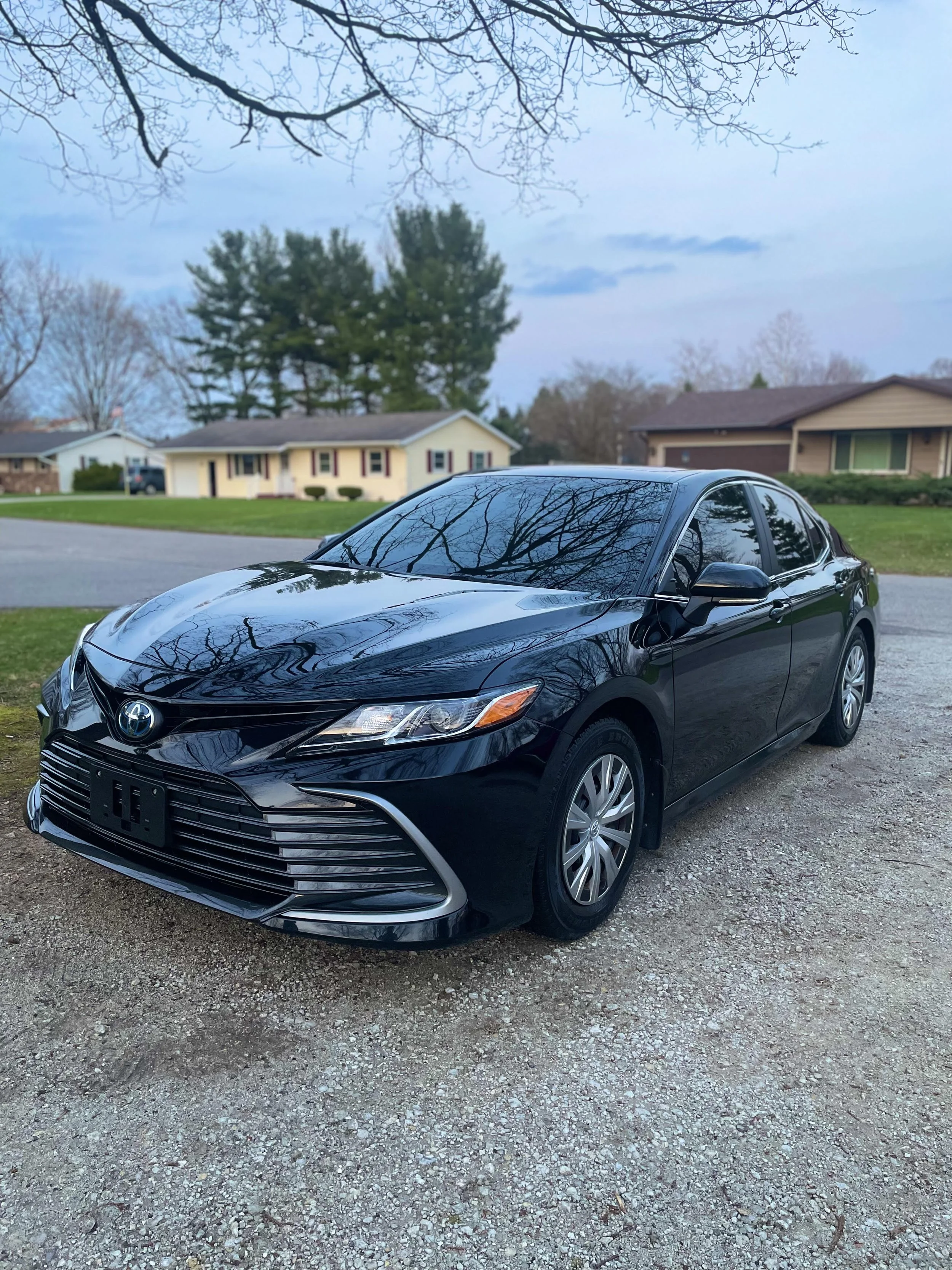 Black Toyota Camry sedan parked on a gravel driveway with leafless trees, suburban houses, and a cloudy sky reflected on its surface.