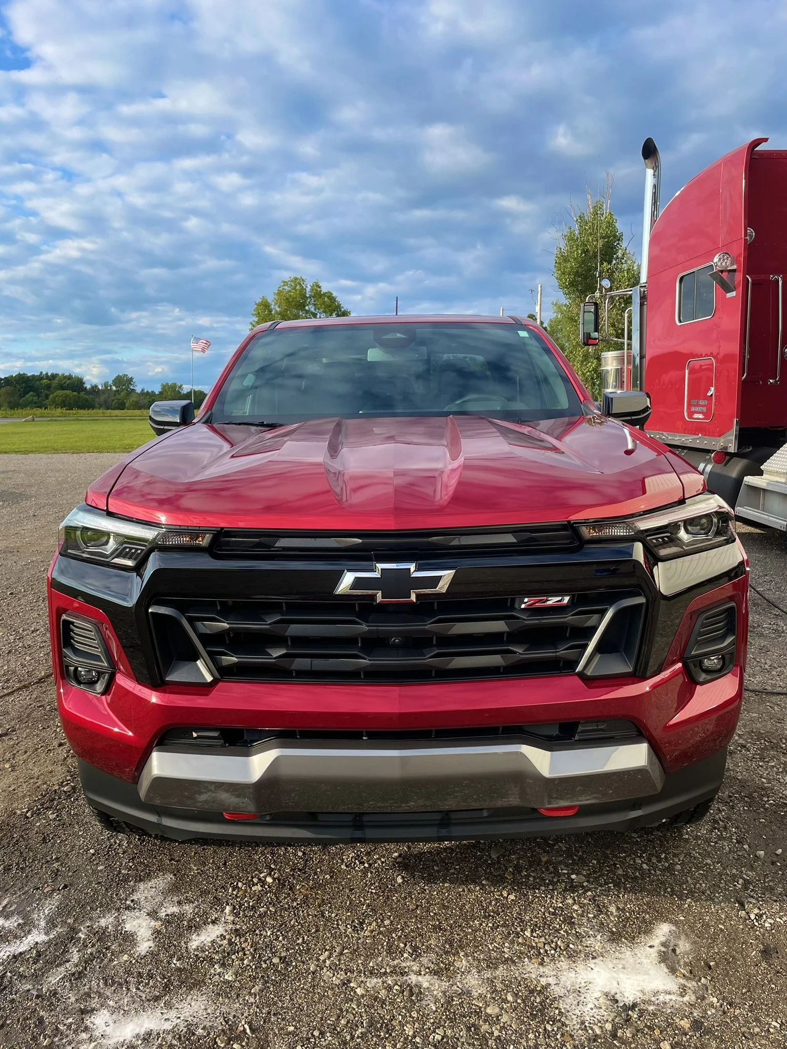 Front view of a red Chevrolet Colorado Z71 pickup truck parked outdoors with a blue sky, clouds, and greenery in the background.