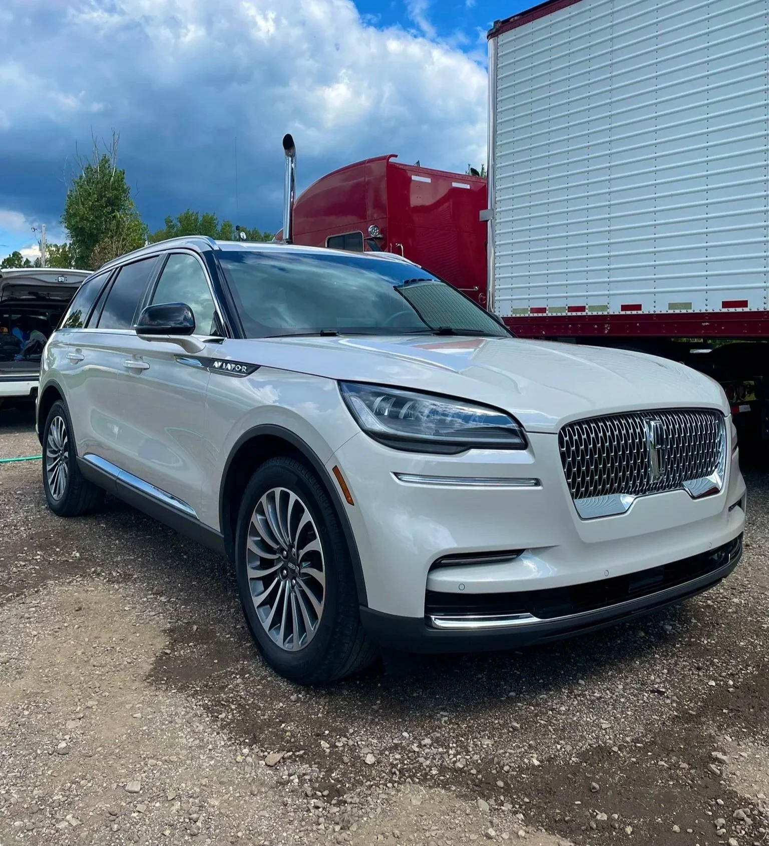 White Lincoln Aviator SUV parked on gravel with semi-truck trailer behind and cloudy sky above.