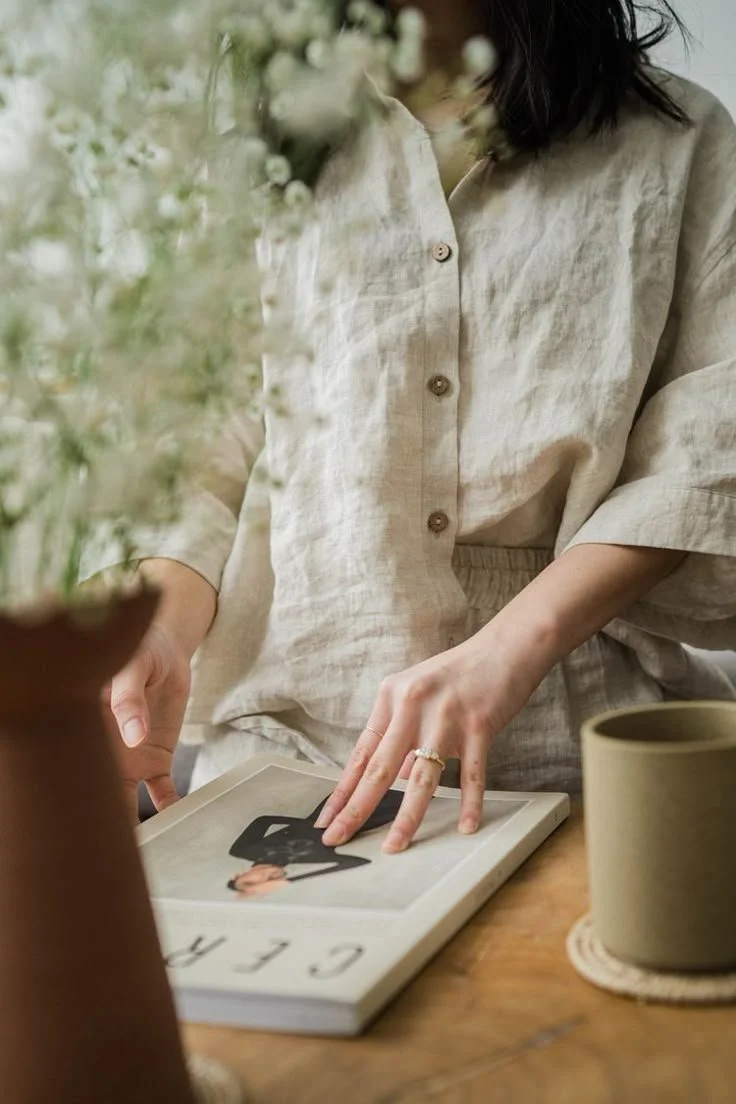 A woman in a beige linen shirt is flipping through a magazine on a wooden table, with a potted plant and a ceramic mug nearby.