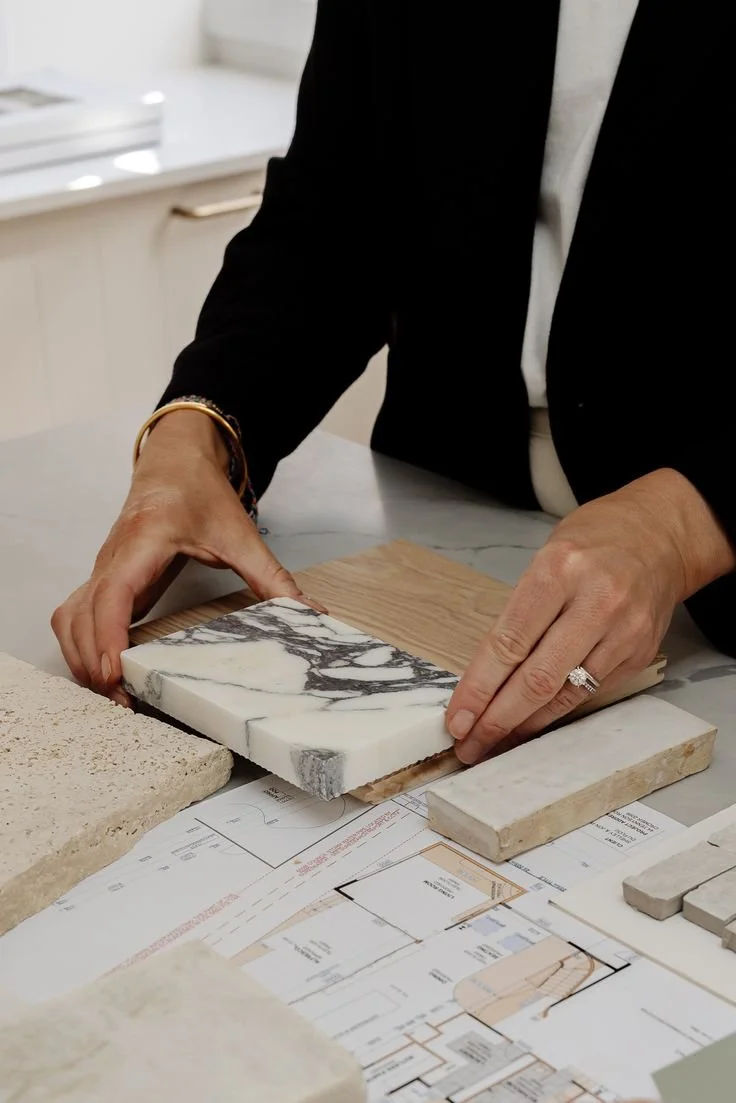 Person in a black blazer and white top is arranging marble and tile samples on a table with architectural plans.