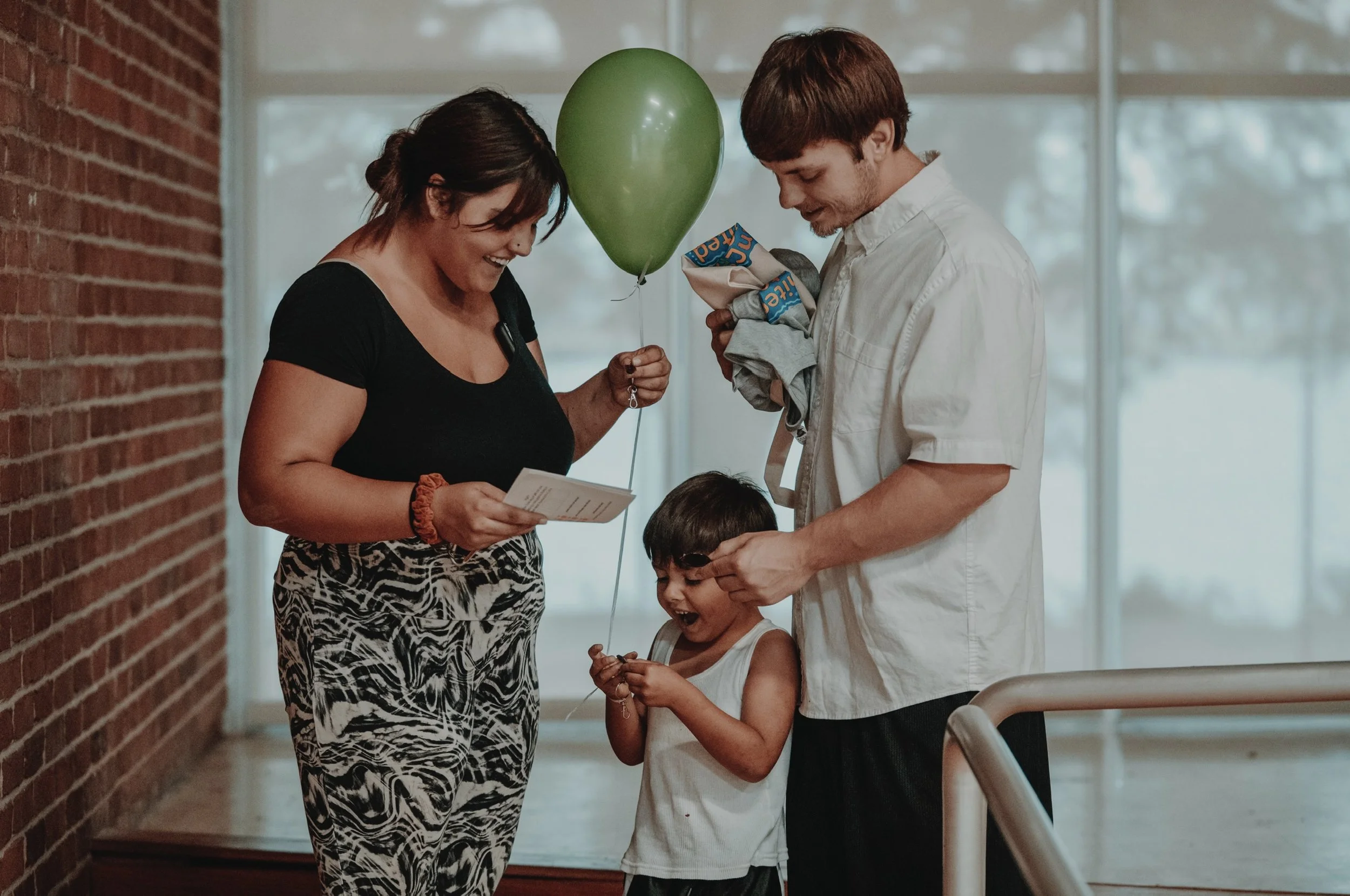 A family celebrating a birthday, with a woman holding a green balloon and a card, a young boy smiling and opening a gift, and a man standing next to him in a room with large windows and a brick wall.