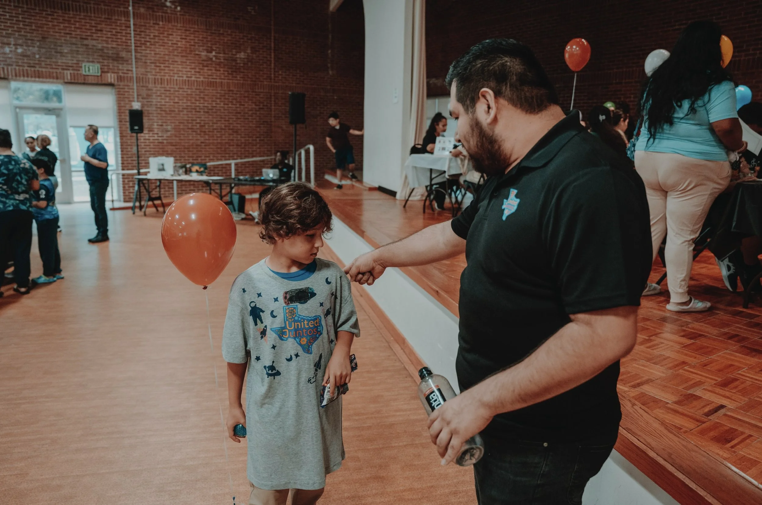 A young boy receives a punch from an adult man at a community event with balloons and themed shirts.