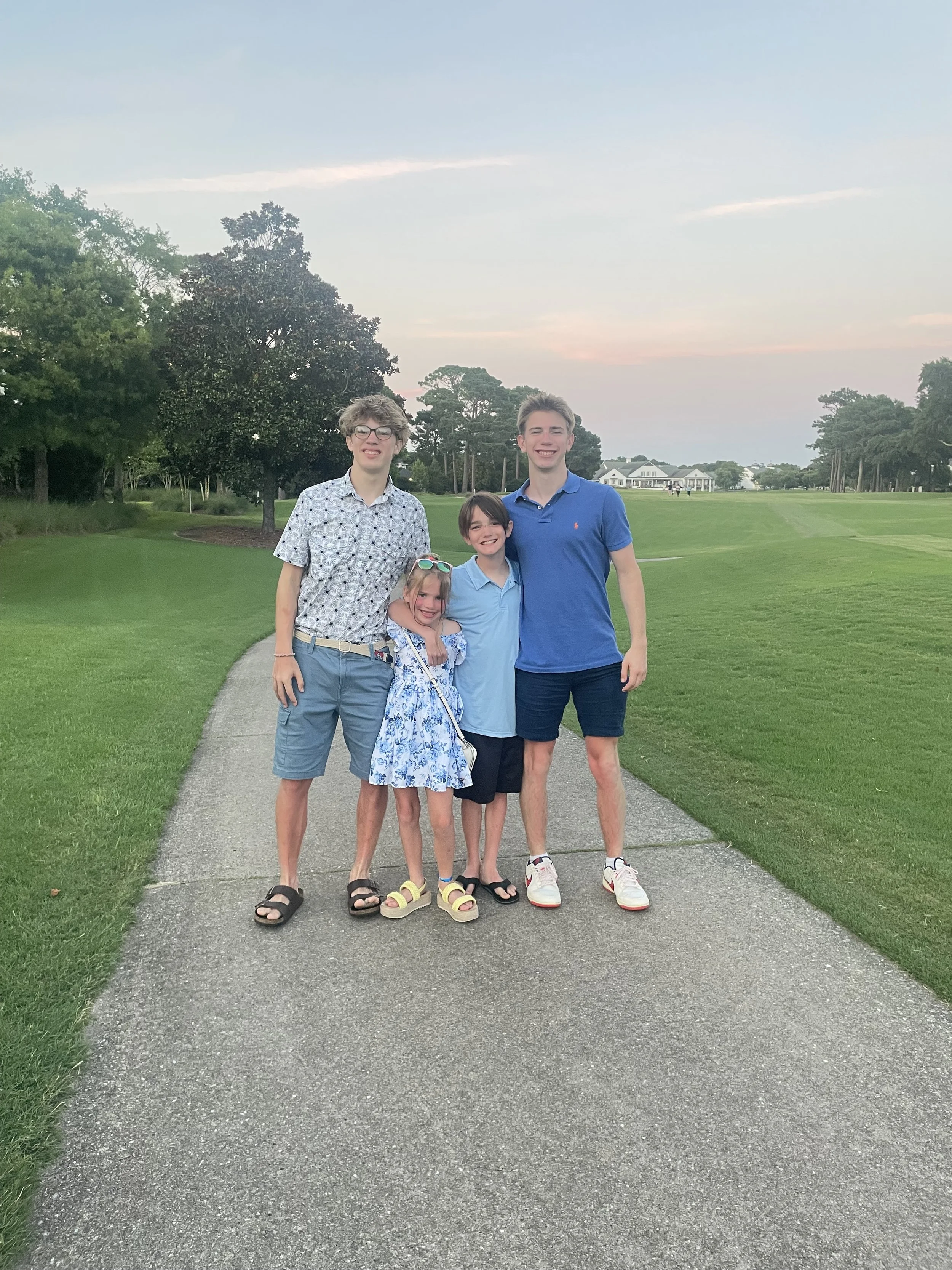 Group of four kids, two boys and two girls, standing on a sidewalk in a park, smiling at the camera with trees and a house in the background, during sunset.