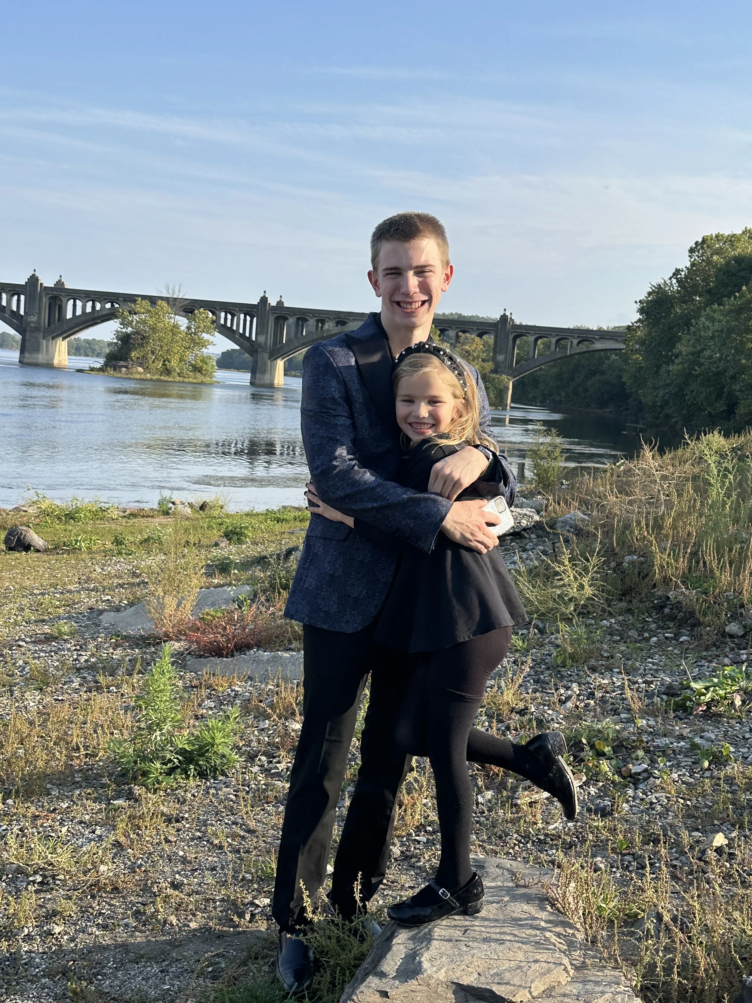 A young man and girl hugging by a river with a bridge in the background, both smiling and dressed in formal clothes.