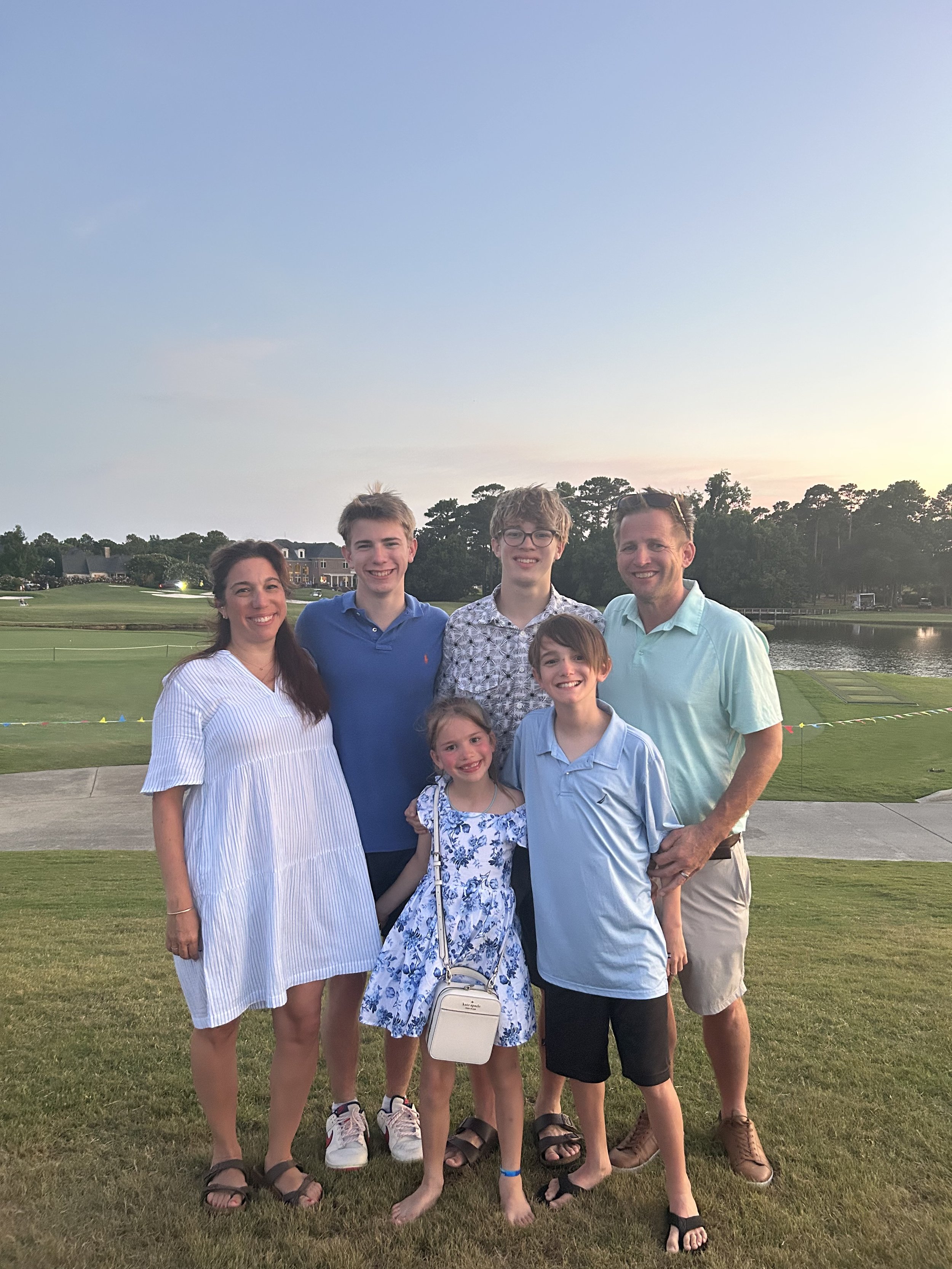 A family of seven standing outdoors on a grassy area near a lake during sunset, smiling at the camera.
