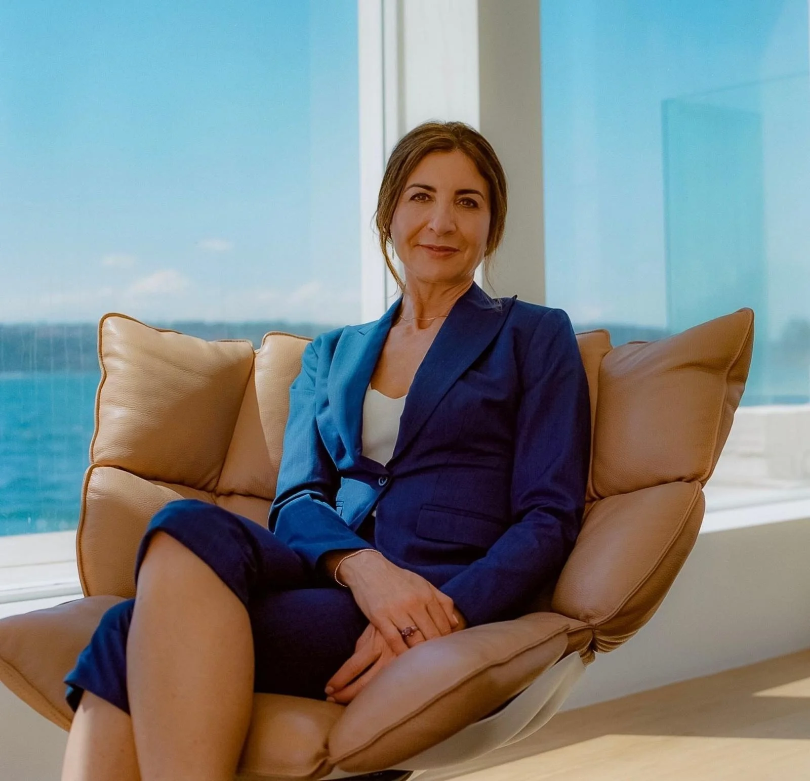 A woman with brown hair wearing a blue blazer and matching pants, sitting on a tan lounge chair with a view of the ocean and sky through large windows.