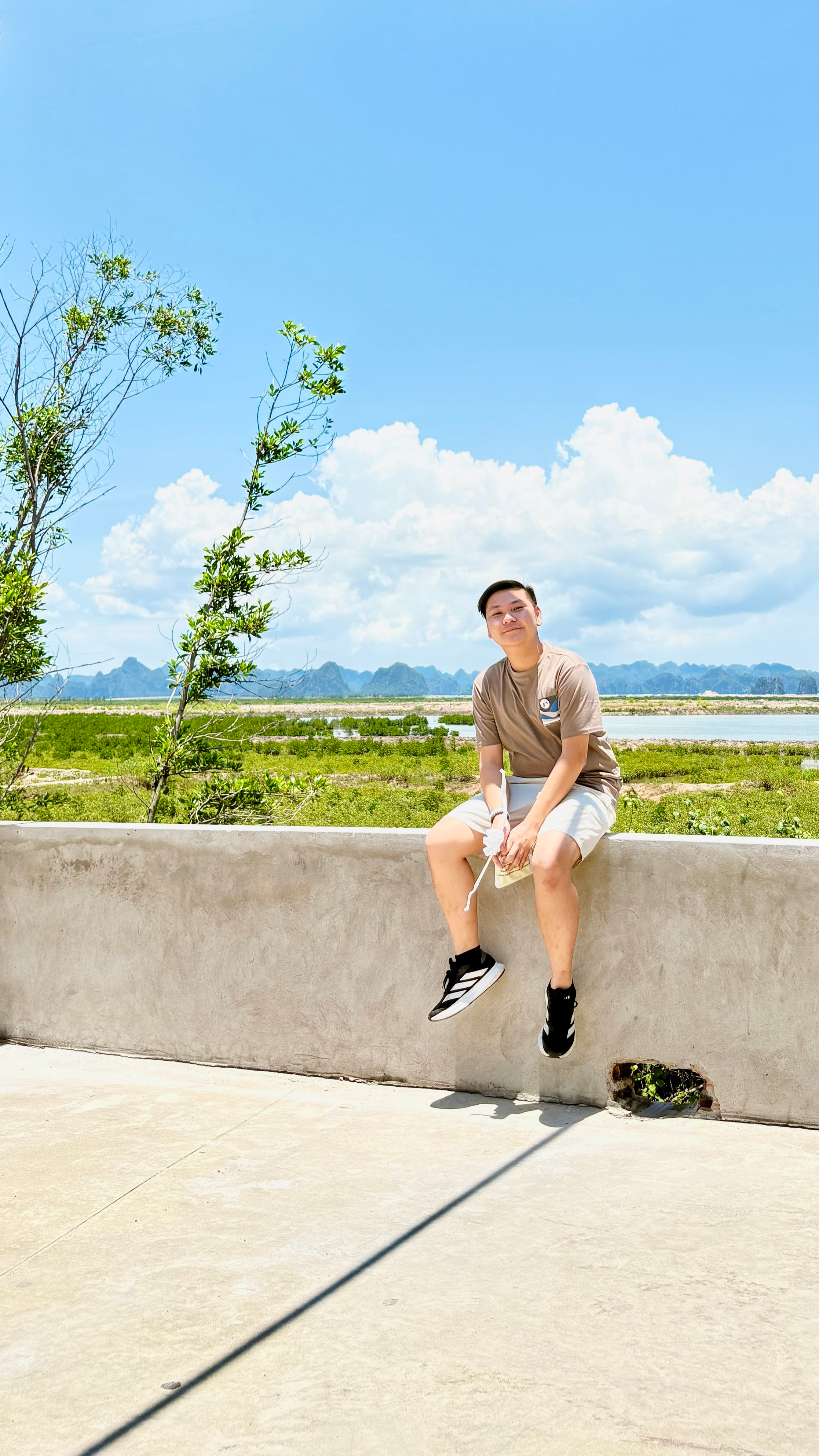 A young man sitting on a concrete wall outdoors, with a scenic landscape of green fields, water, and mountains in the background under a blue sky with clouds.