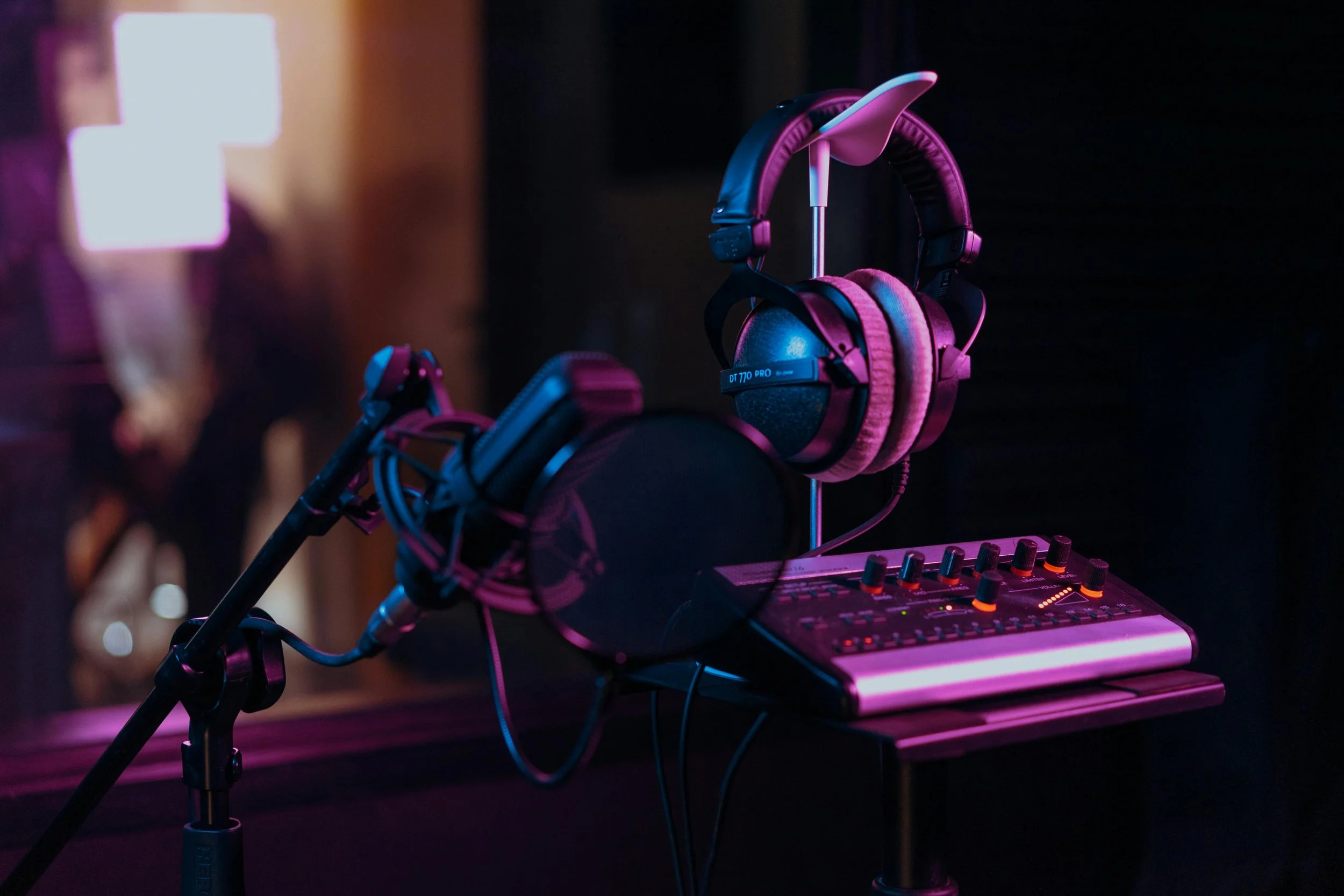 DJ headphones, microphone, and audio mixer on a stand in a dimly lit studio or stage with purple and pink lighting.