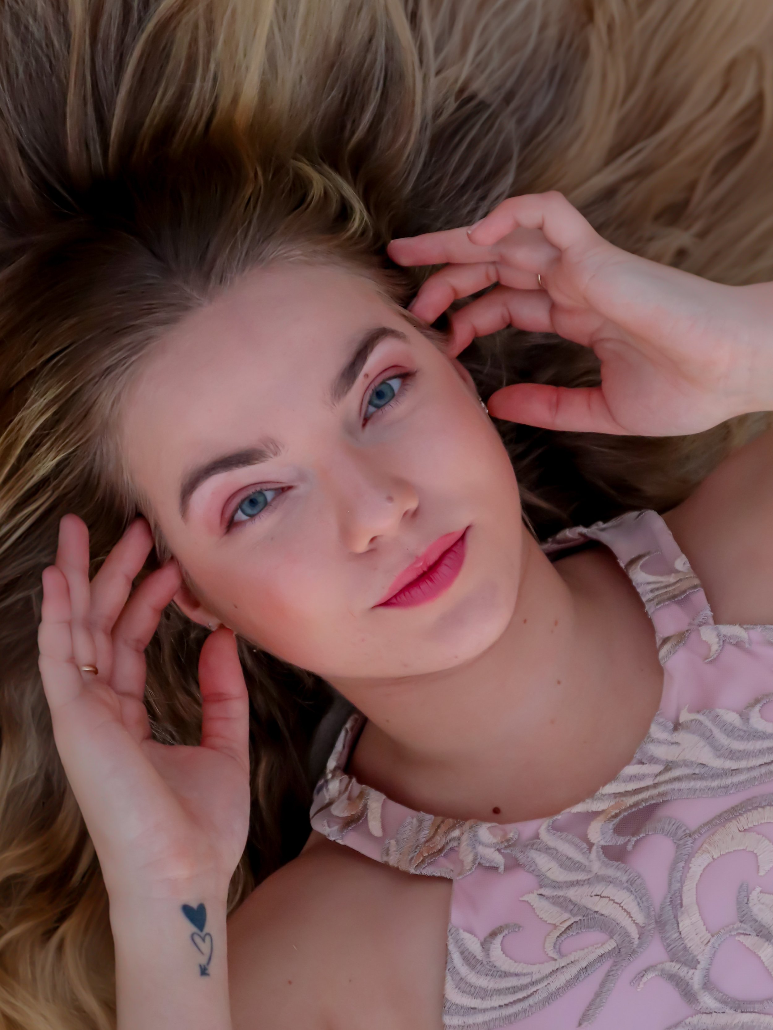 A young woman with blonde hair, blue eyes, and light skin lying on a wooden surface, looking at the camera with a slight smile, wearing a pink lace dress, with a small tattoo on her left wrist.