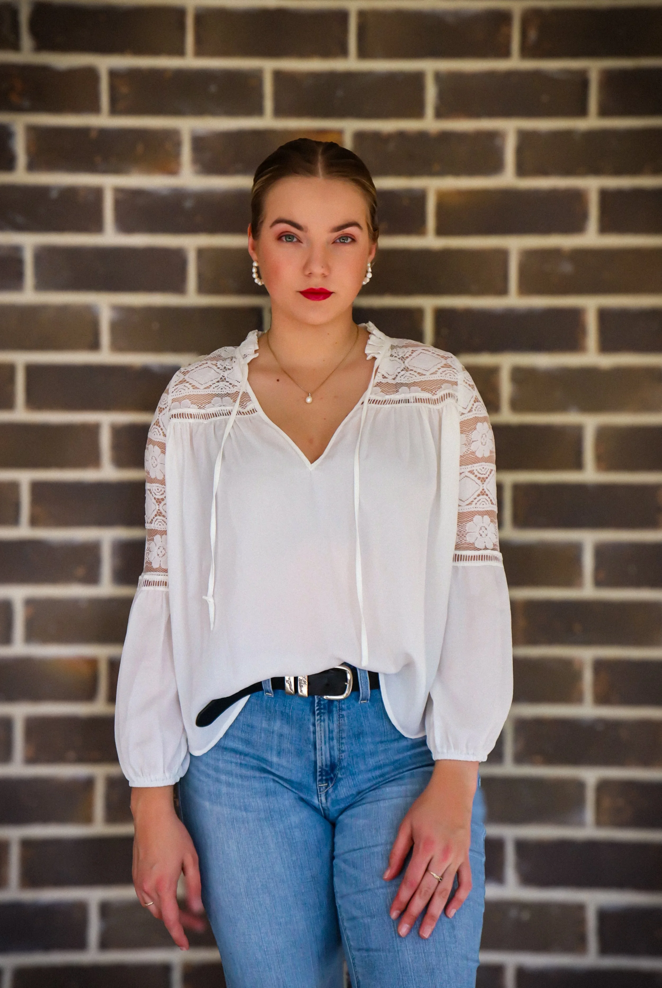A young woman with blonde hair, red lipstick, and pearl earrings standing in front of a brick wall, wearing a white lace blouse and blue jeans.