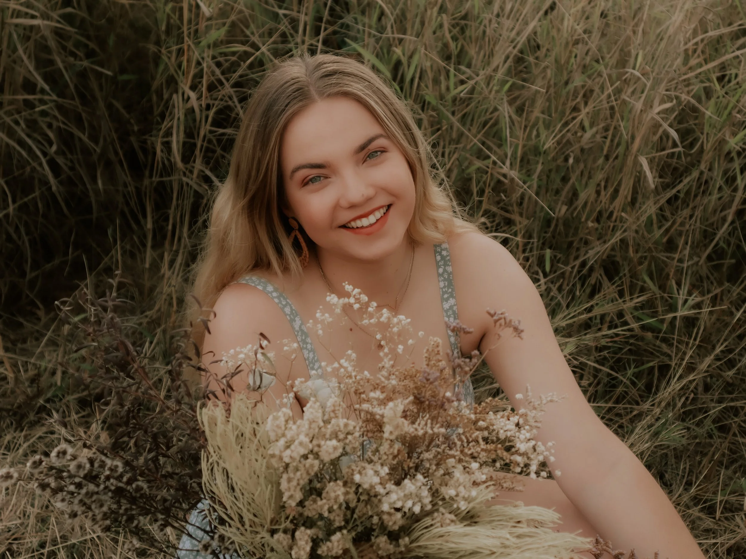 A smiling woman with blonde hair, wearing a sleeveless dress with patterned straps, sitting in tall grass with a bouquet of dried flowers in front of her.
