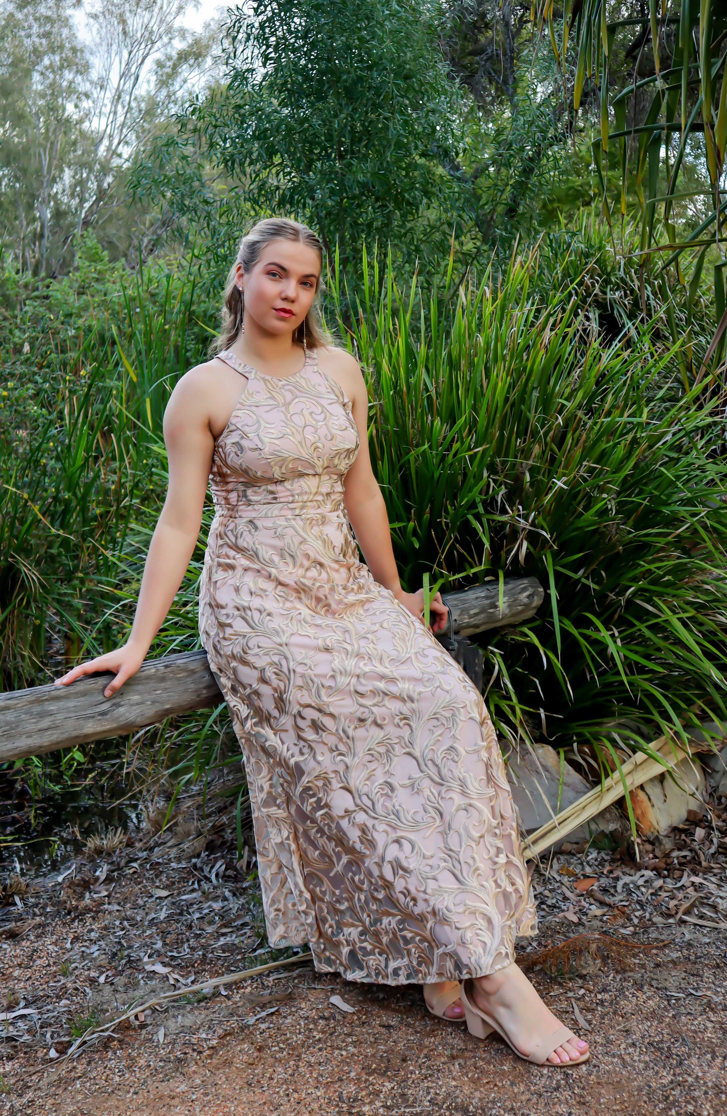 Young woman in a patterned dress sitting on a wooden railing surrounded by green foliage in an outdoor natural setting.
