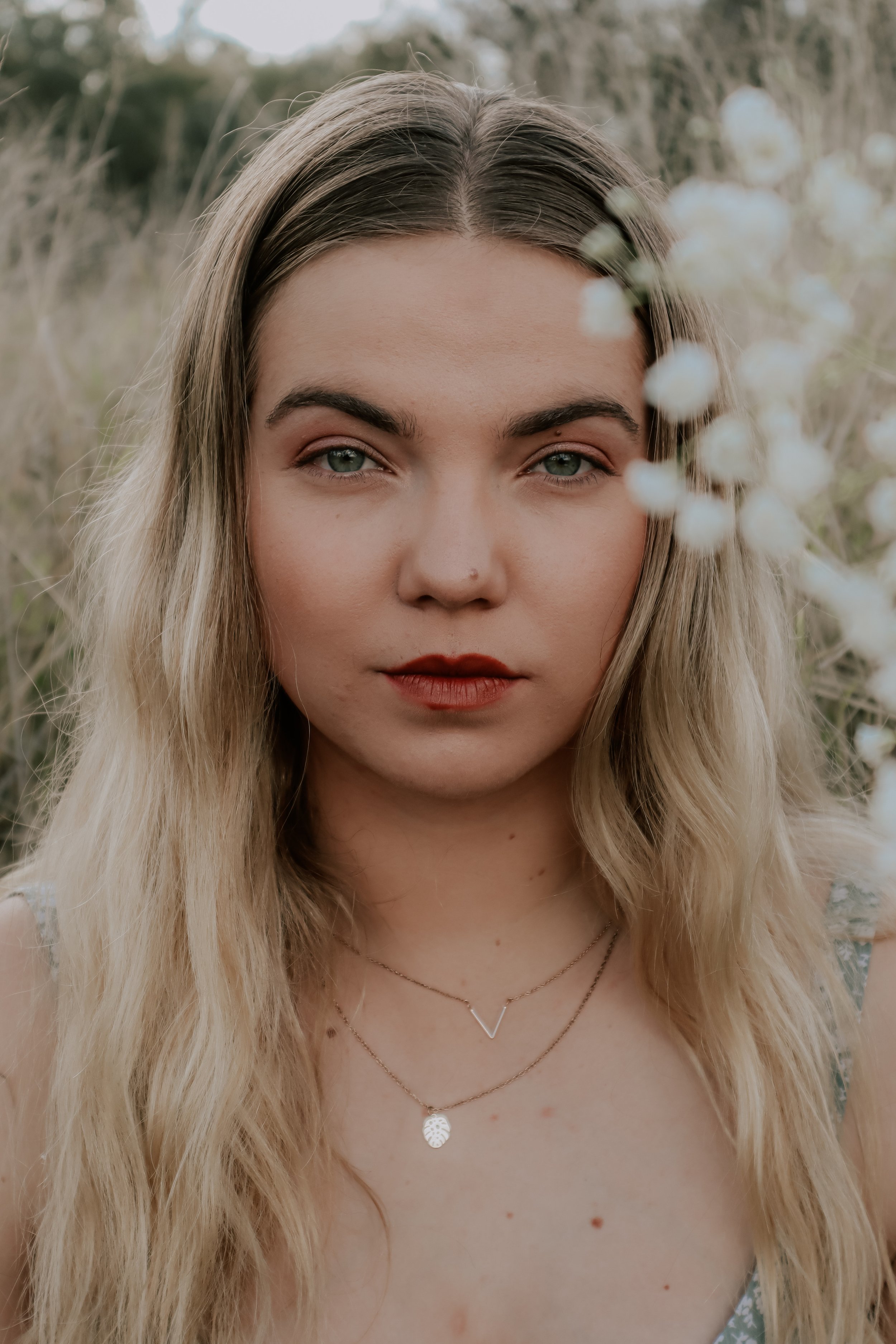 Close-up portrait of a young woman with blonde wavy hair, piercing blue eyes, wearing red lipstick, layered necklaces, and a floral dress in a natural outdoor setting with blurred white flowers in the foreground.