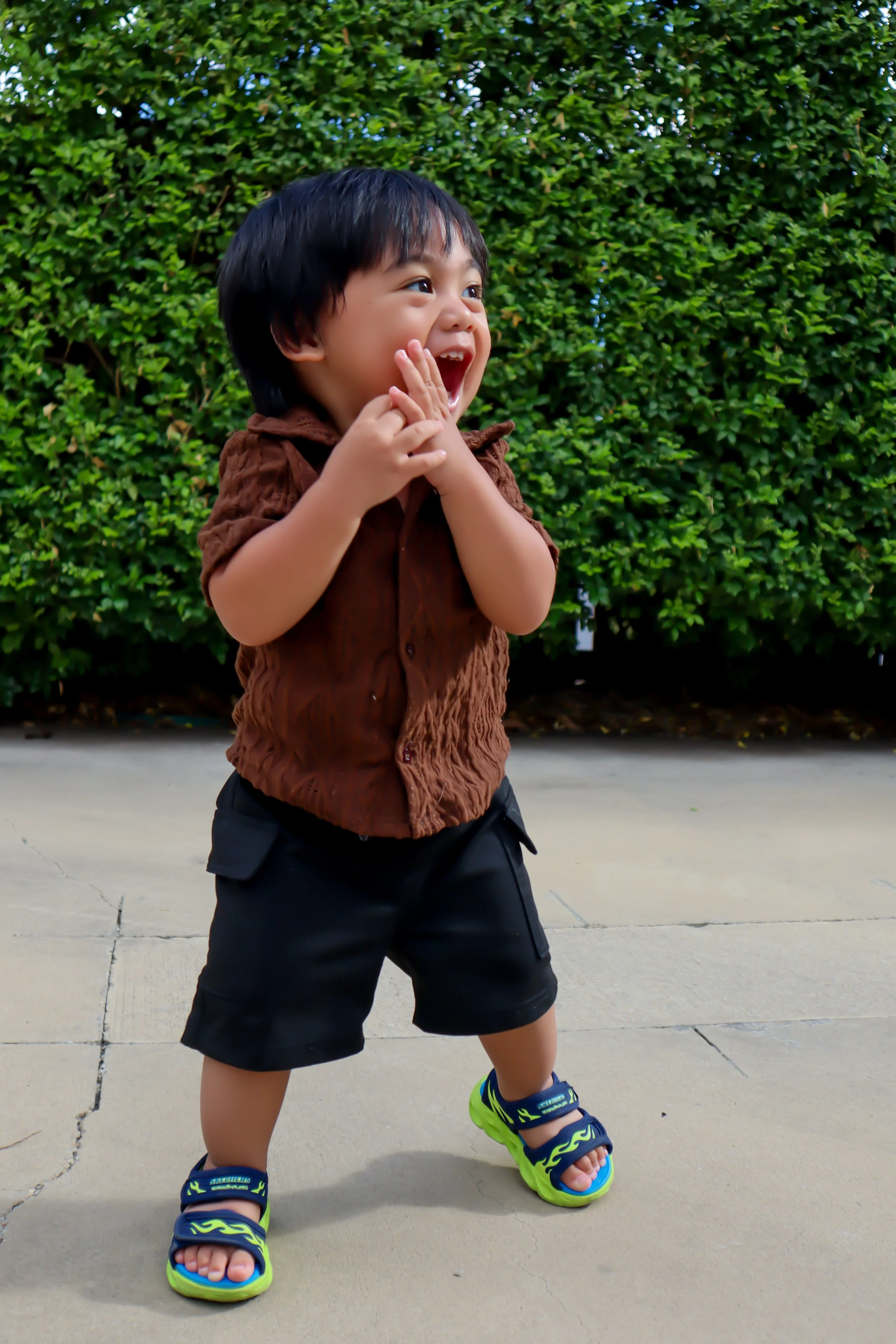 A young boy with black hair and light skin laughing outdoors on a gray sidewalk, wearing a brown shirt, black shorts, and colorful blue and green sandals, with green bushes in the background.