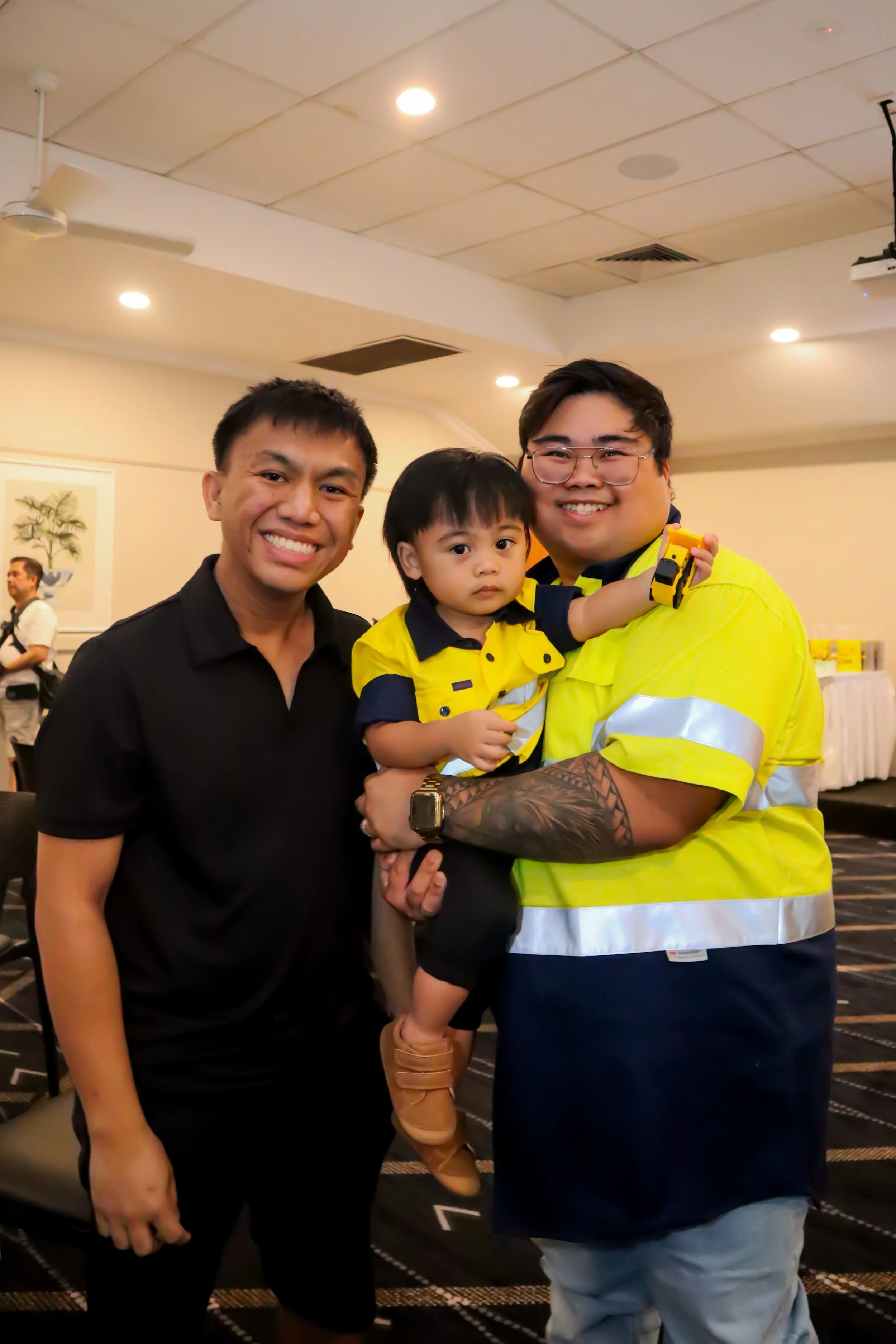 Three people, two men and a young boy, smiling and posing indoors. One man is wearing a black polo shirt, the other is in a reflective yellow safety shirt holding the boy, who is dressed in a yellow and navy shirt and tan shoes.