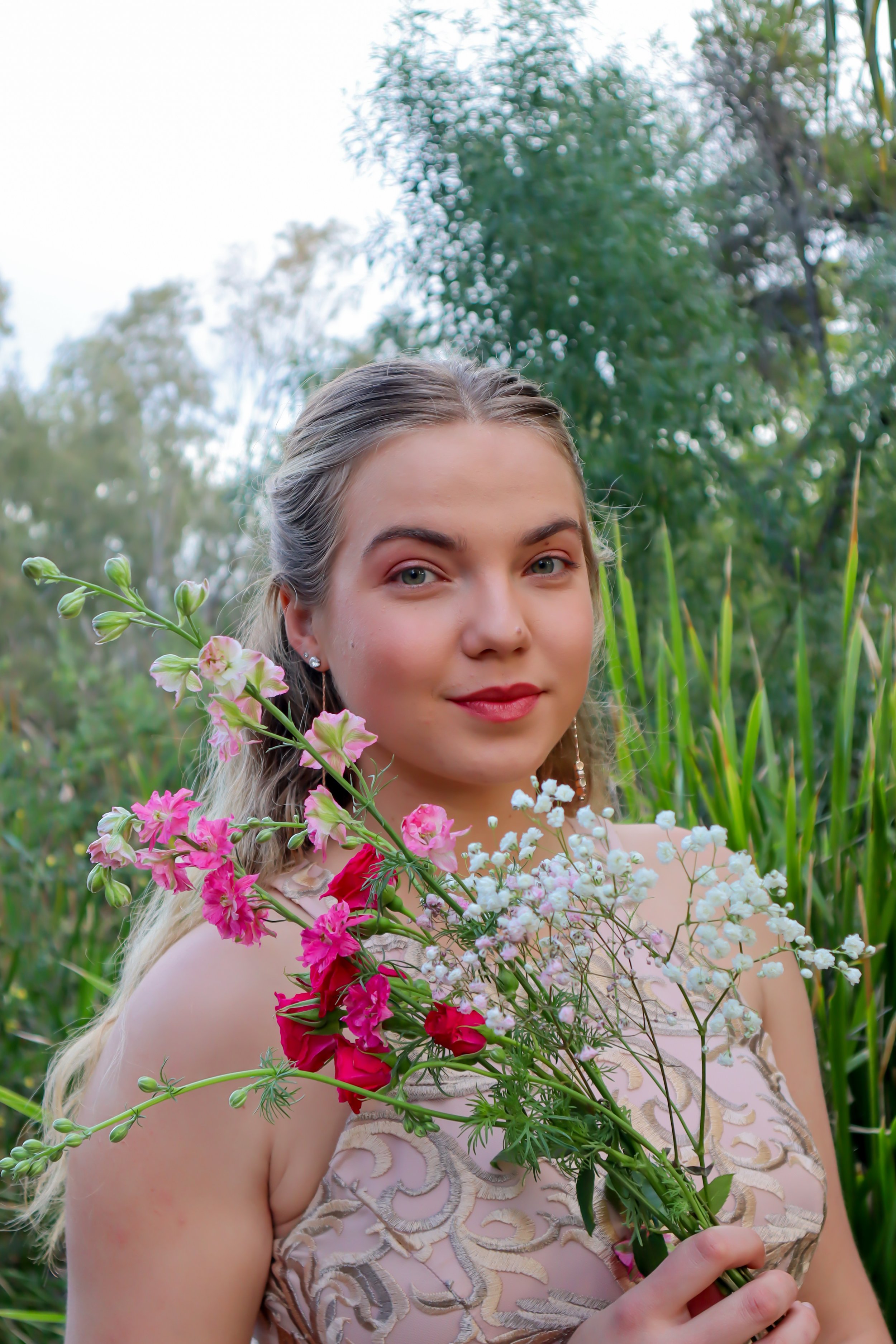 A woman with blonde hair holding a bouquet of pink, white, and red flowers outdoors, with green trees and tall grass in the background.