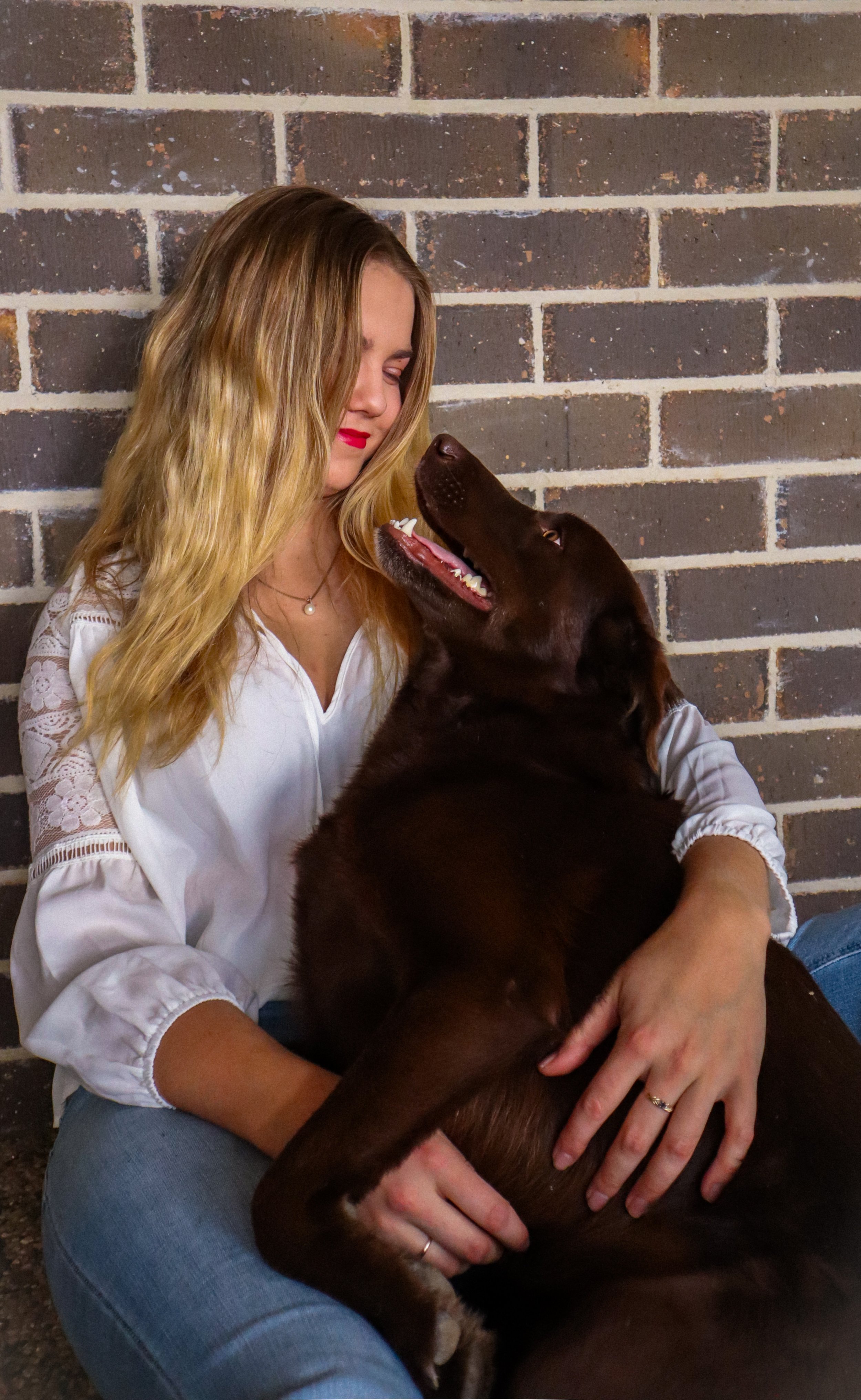 A woman with long blonde hair and red lipstick sitting against a brick wall, embracing and smiling at a large brown dog that is looking up at her with its mouth open.