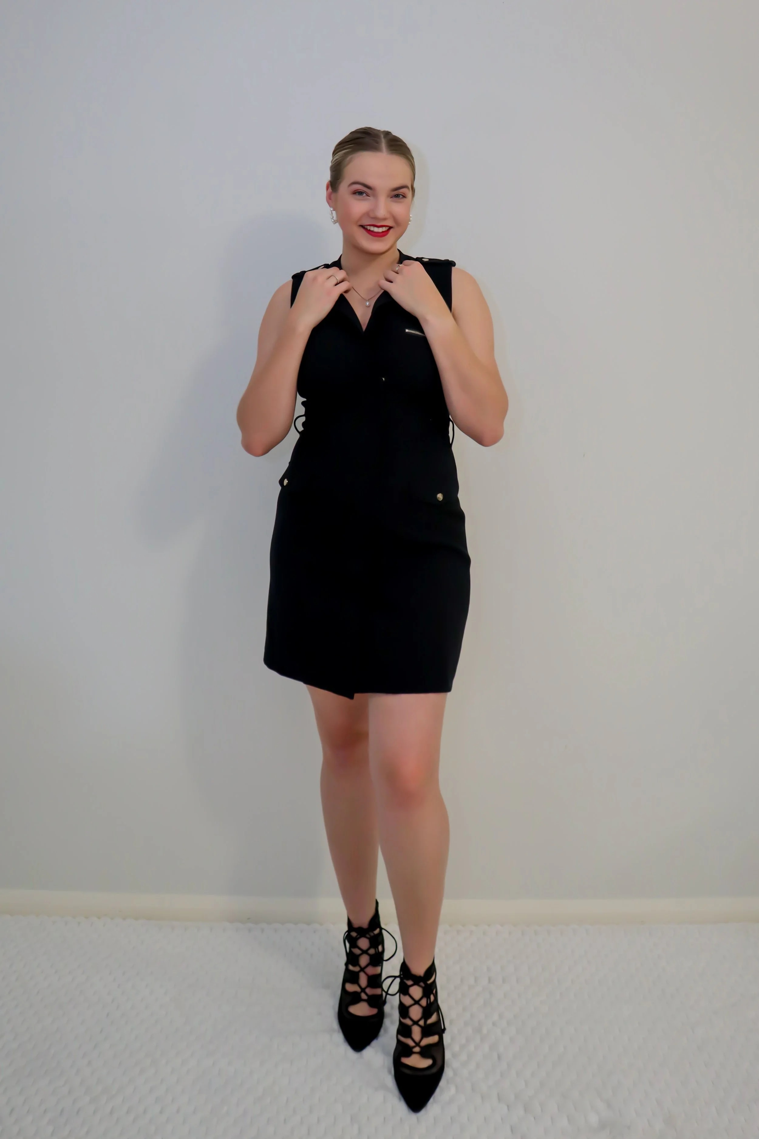 A young woman with blonde hair, dressed in a black sleeveless dress and black high-heeled shoes with laces, standing against a plain white wall and smiling.