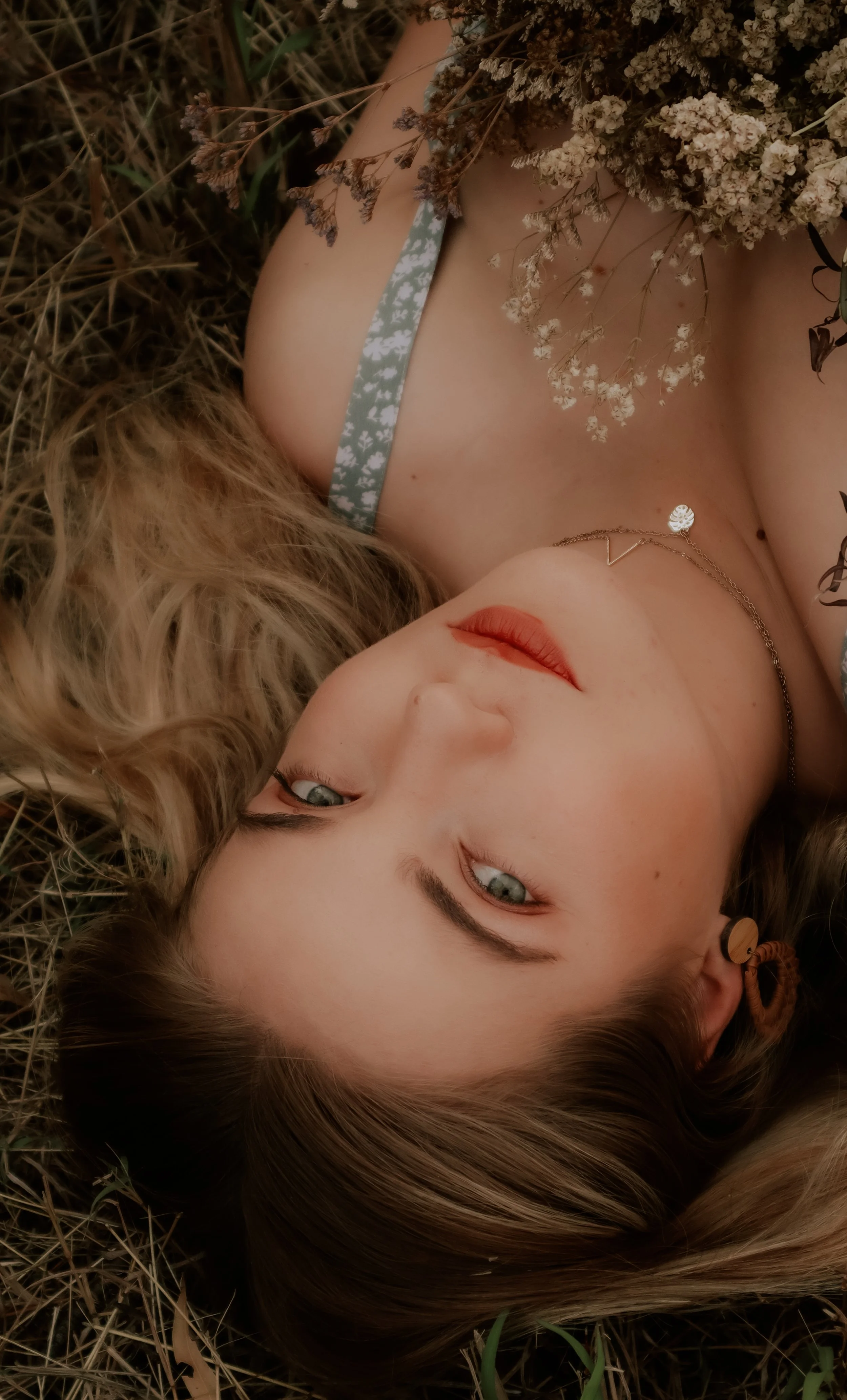 A woman with blonde hair and blue eyes lying on the ground surrounded by dry grass, holding a bouquet of dried flowers, wearing a floral dress strap and a necklace.