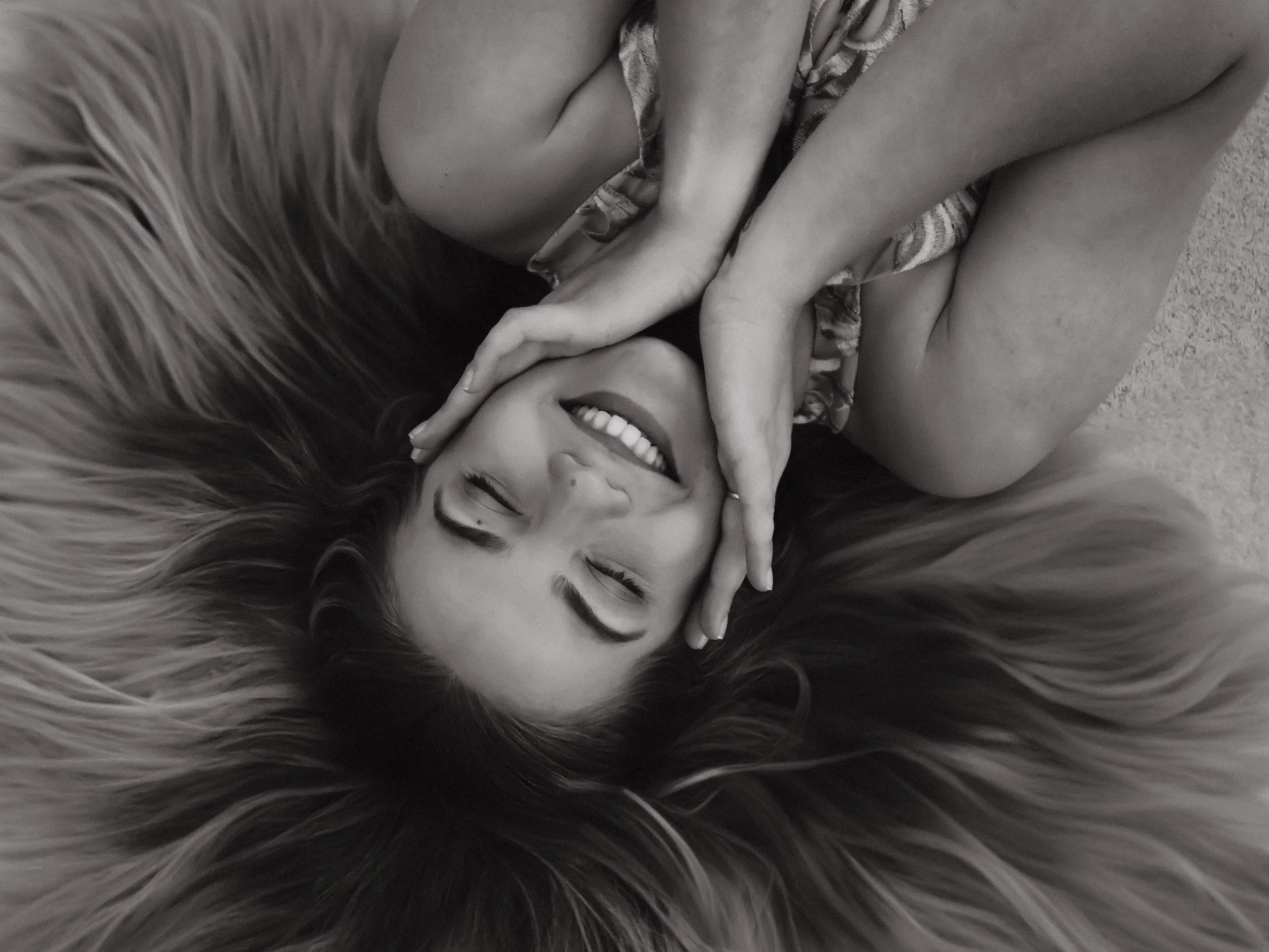 A woman lying on a furry surface, smiling with eyes closed, hands framing her face.