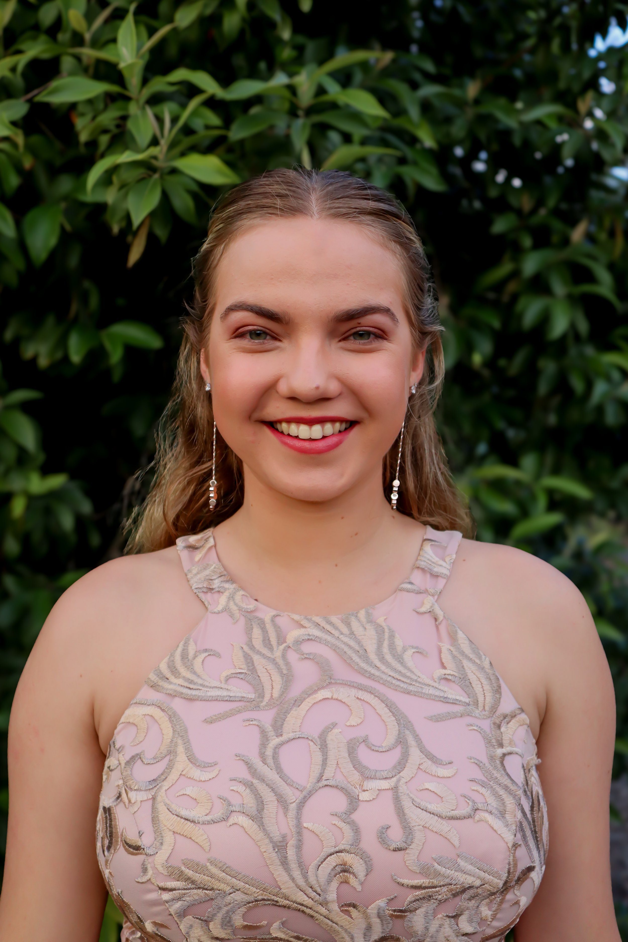 A young woman with wavy blonde hair and jewelry, smiling outdoors in front of green leafy bushes, wearing a pink and beige embroidered top.