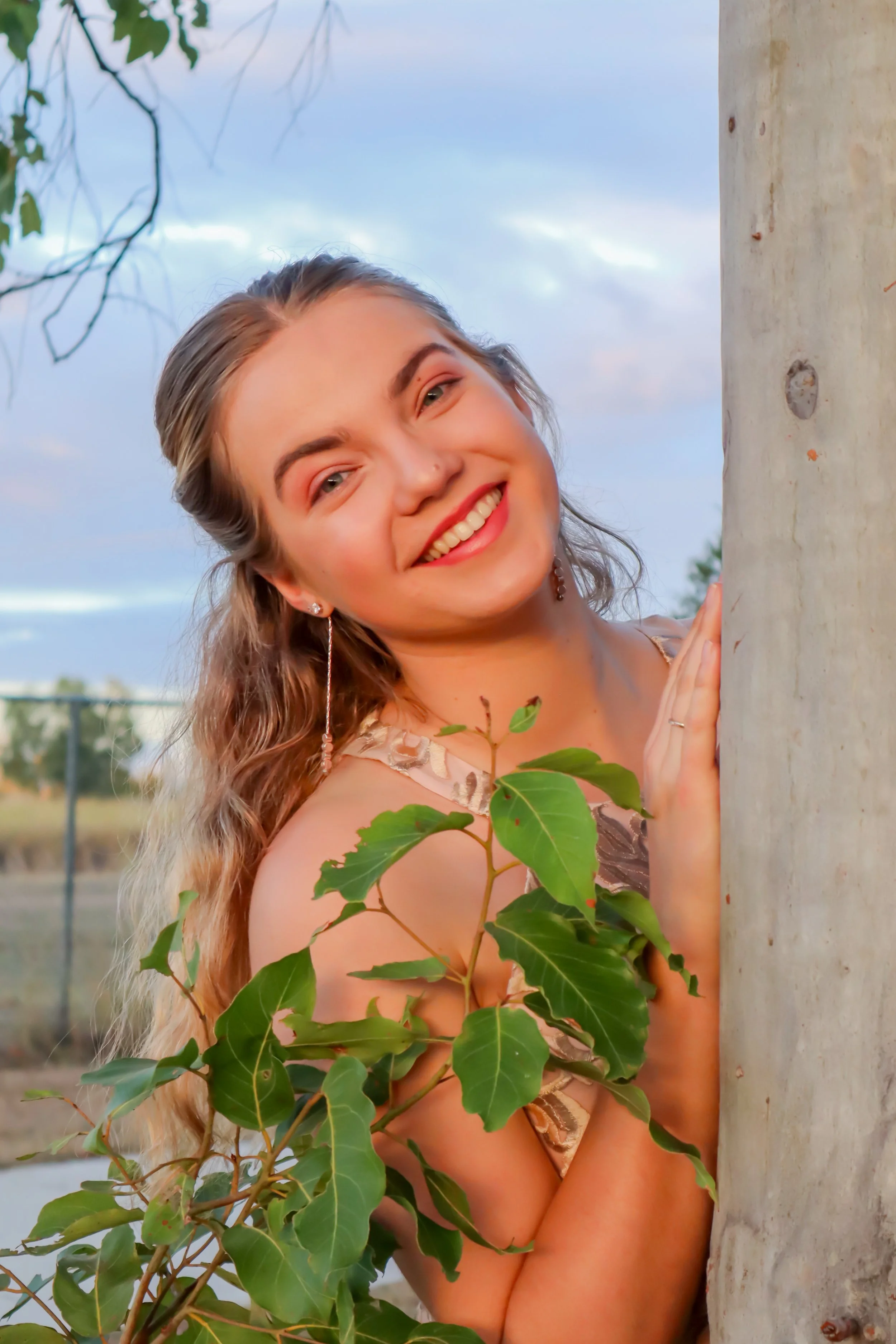 A young woman with wavy blonde hair, smiling and peeking out from behind a tree trunk, outdoors during sunset, with green leaves and a cloudy sky in the background.