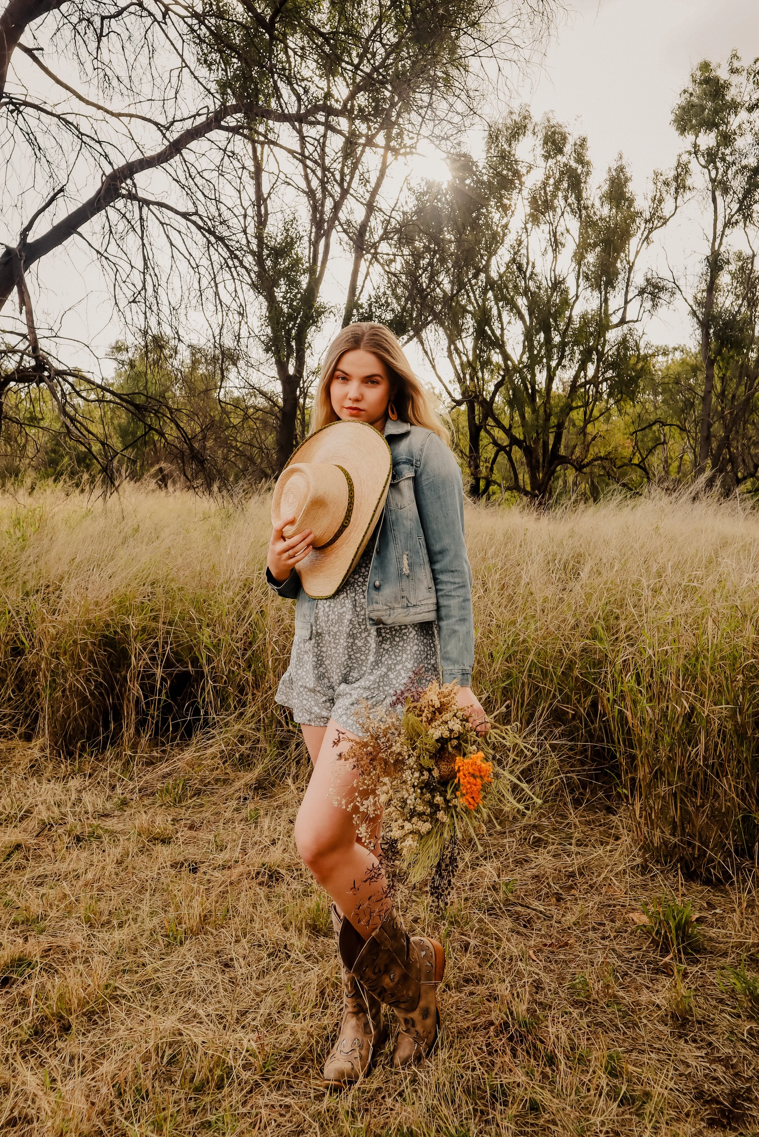 A young woman in a field holding a bouquet of wildflowers and a straw hat, wearing brown cowboy boots, denim jacket, and floral shorts with a natural background of trees and grass.