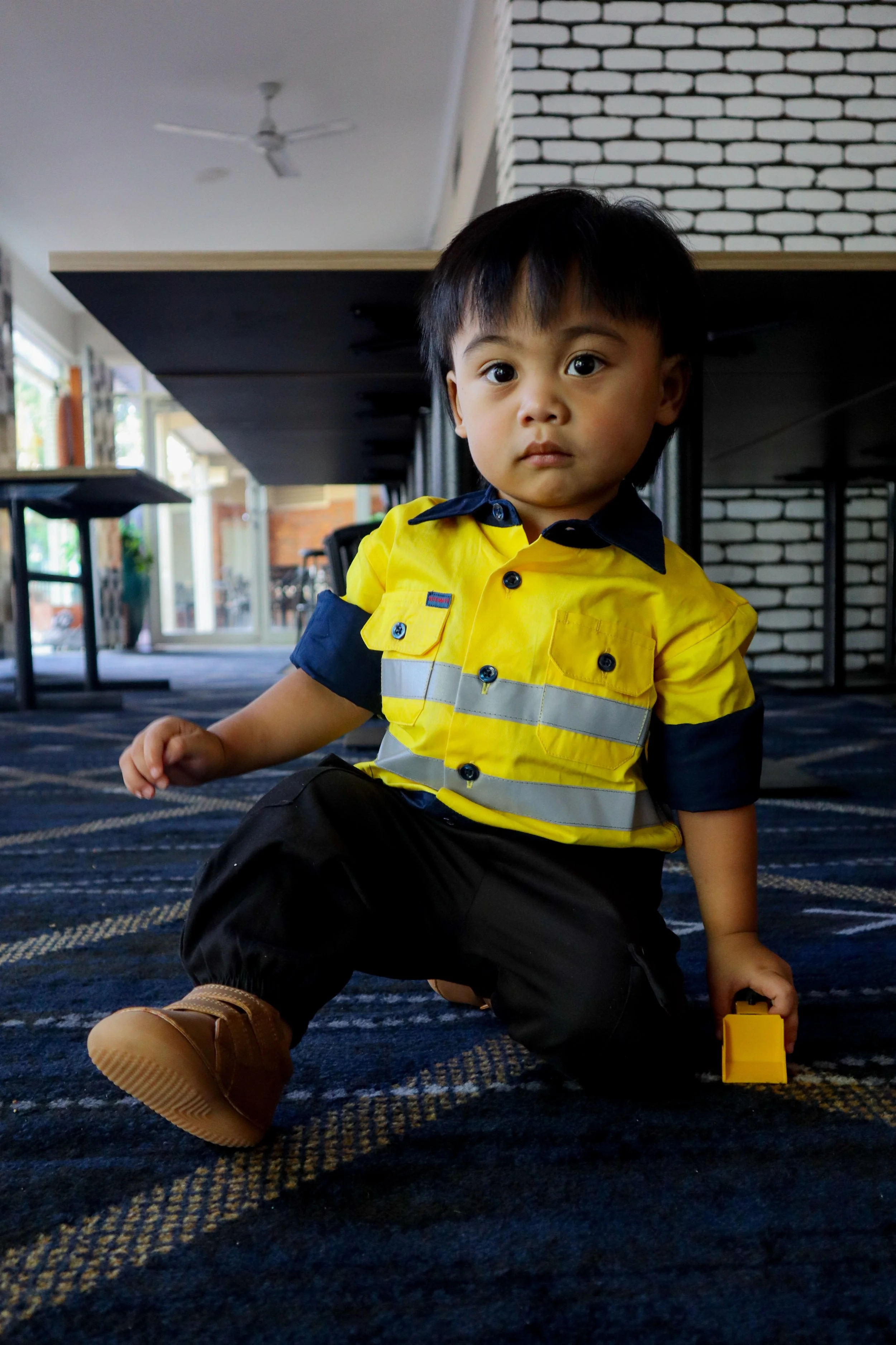 Young child dressed in a yellow and navy blue reflective safety vest, black pants, and tan boots, kneeling on a patterned carpet inside a building with large windows and a brick wall in the background.
