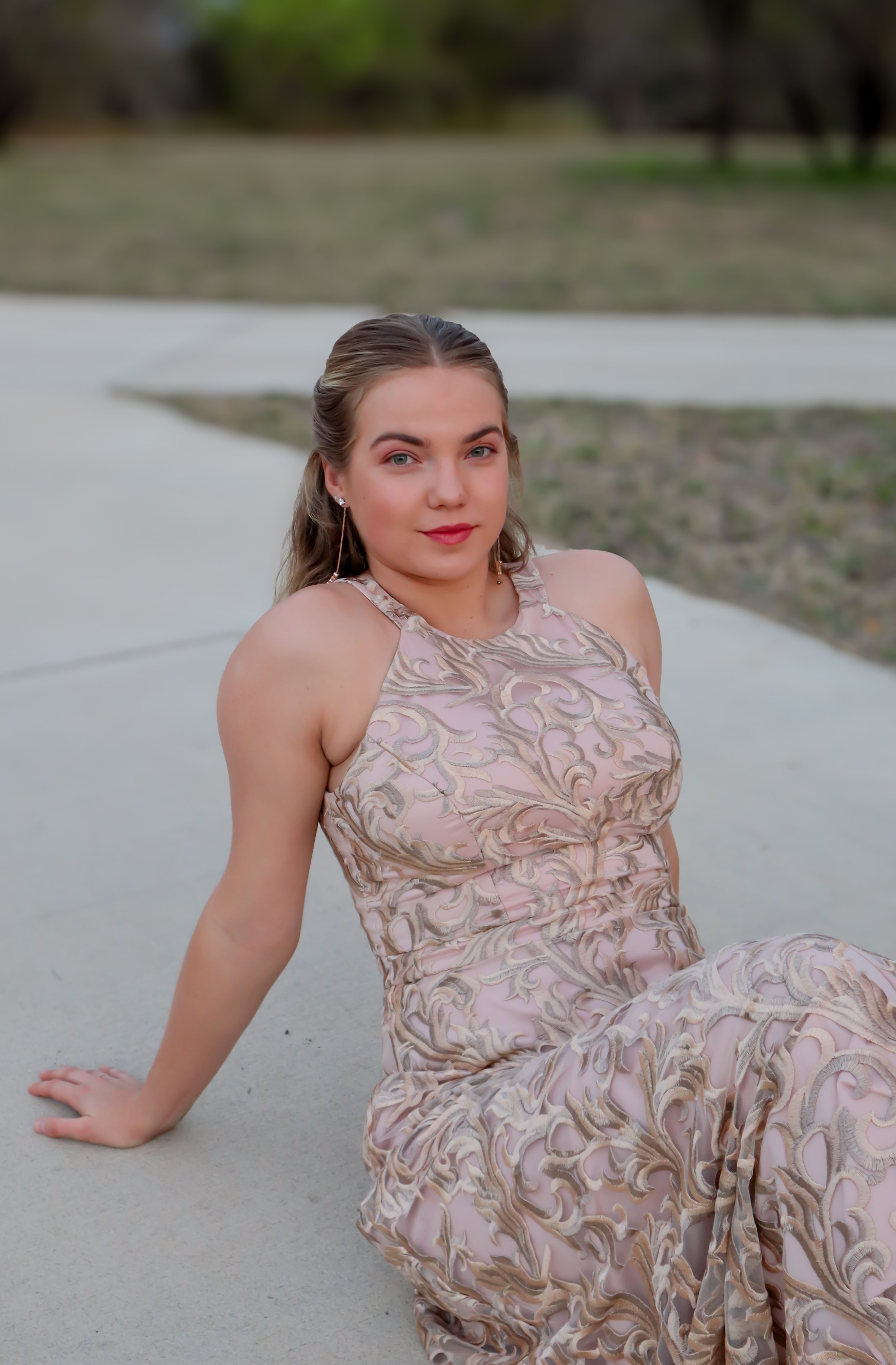 A young woman in a pink and gold patterned dress sitting on a concrete surface outdoors, with grass and trees in the background.