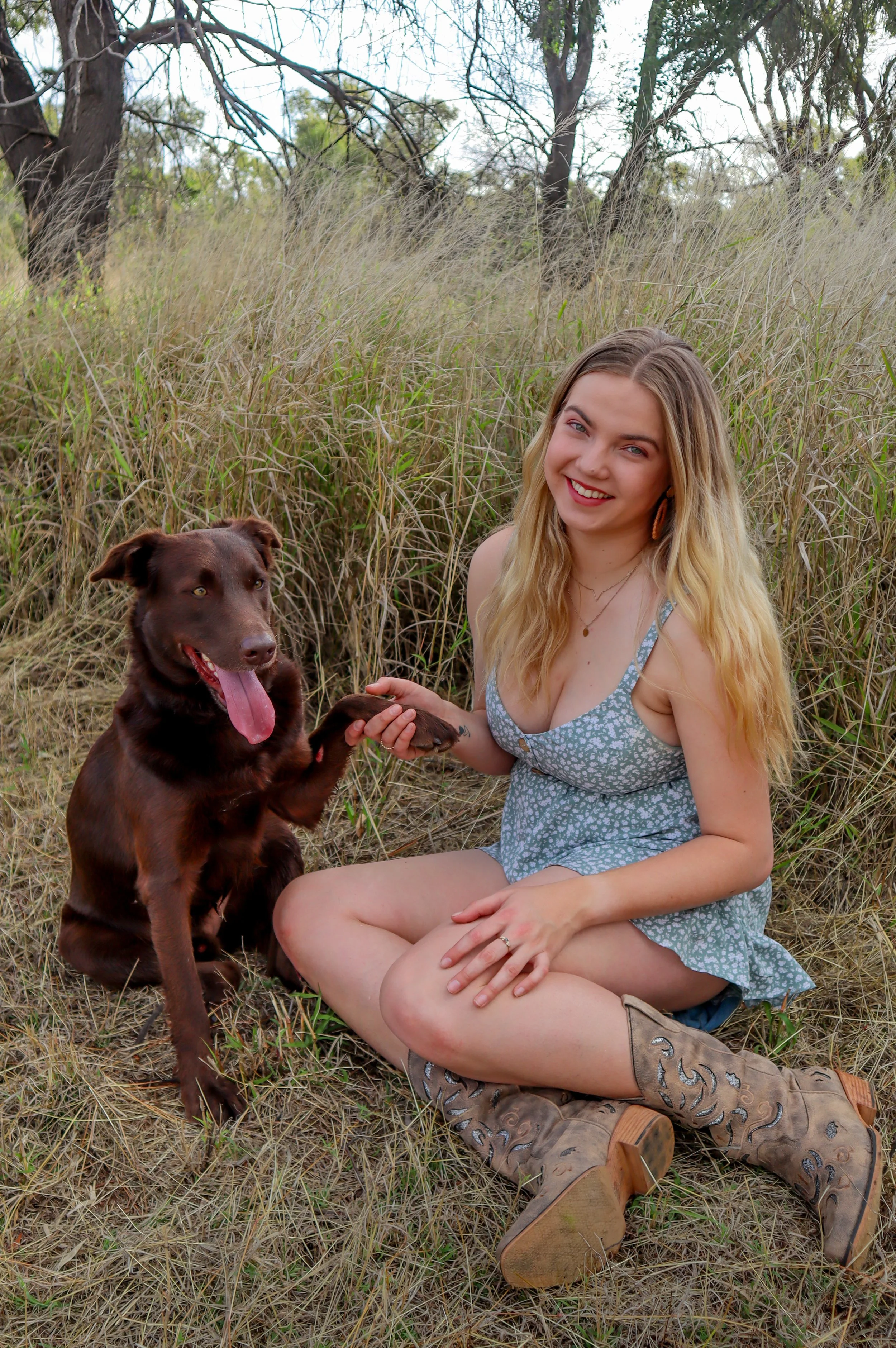A woman with long blonde hair sitting on the grass in a field holding hands with a brown dog, both smiling at the camera.