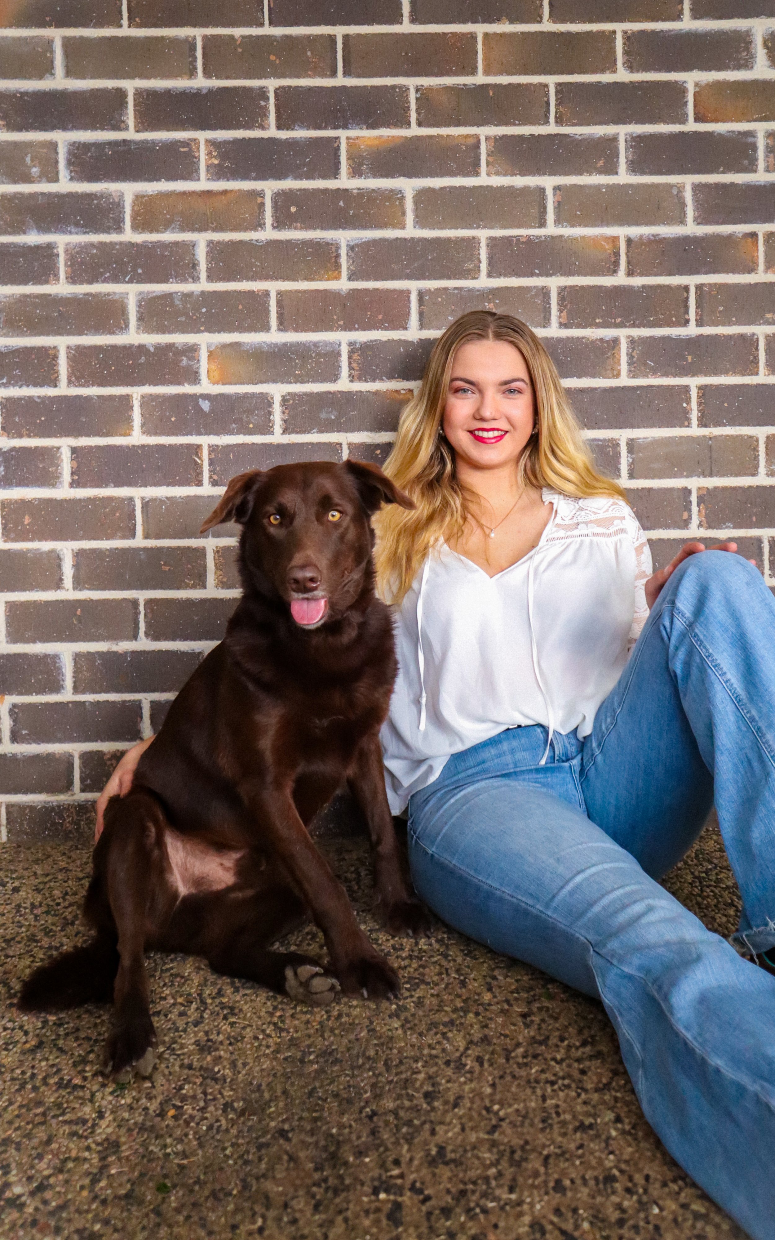 A young woman with blonde hair and red lipstick sitting on the ground next to a brown dog with yellow eyes, both leaning against a brick wall.