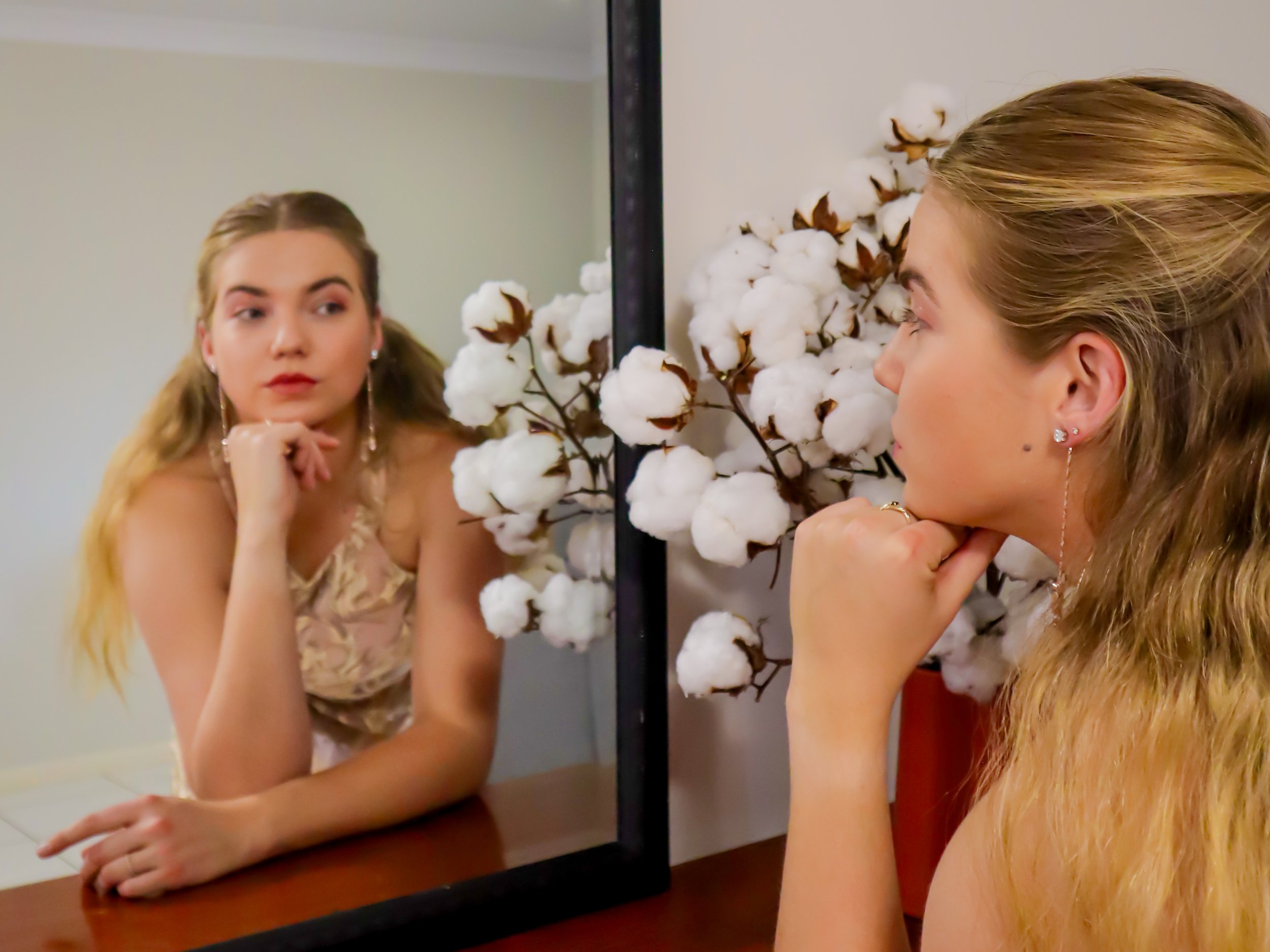 A young woman looking at her reflection in a mirror, resting her chin on her hand, with a cotton plant in front of her. She has long blonde hair, earrings, and is wearing a sleeveless top.