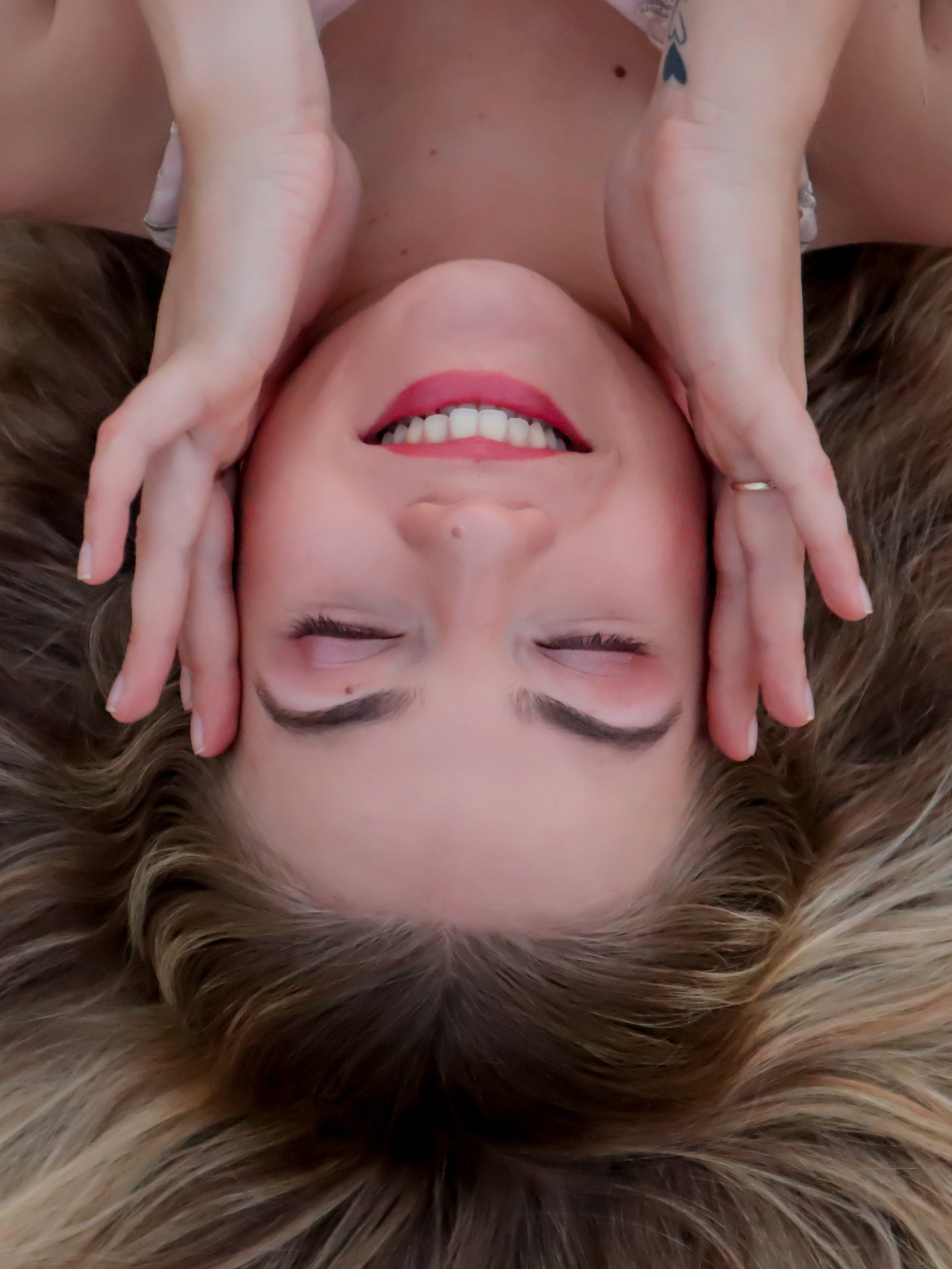 A woman with long hair lying down, smiling with her eyes closed, holding her face with her hands.