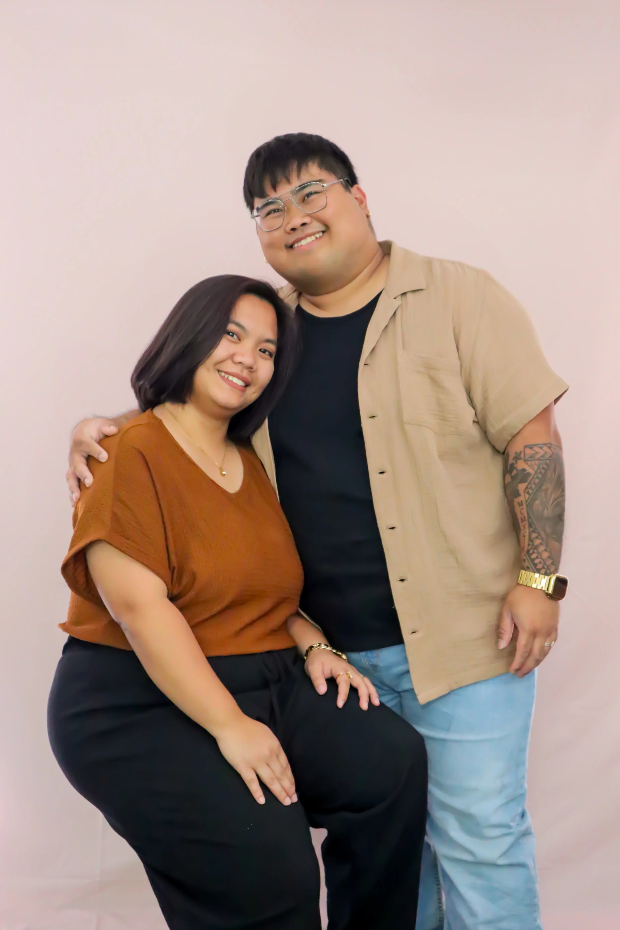 A smiling couple stands close together in front of a light pink background, with the woman seated and the man standing behind her with his arm around her shoulder.