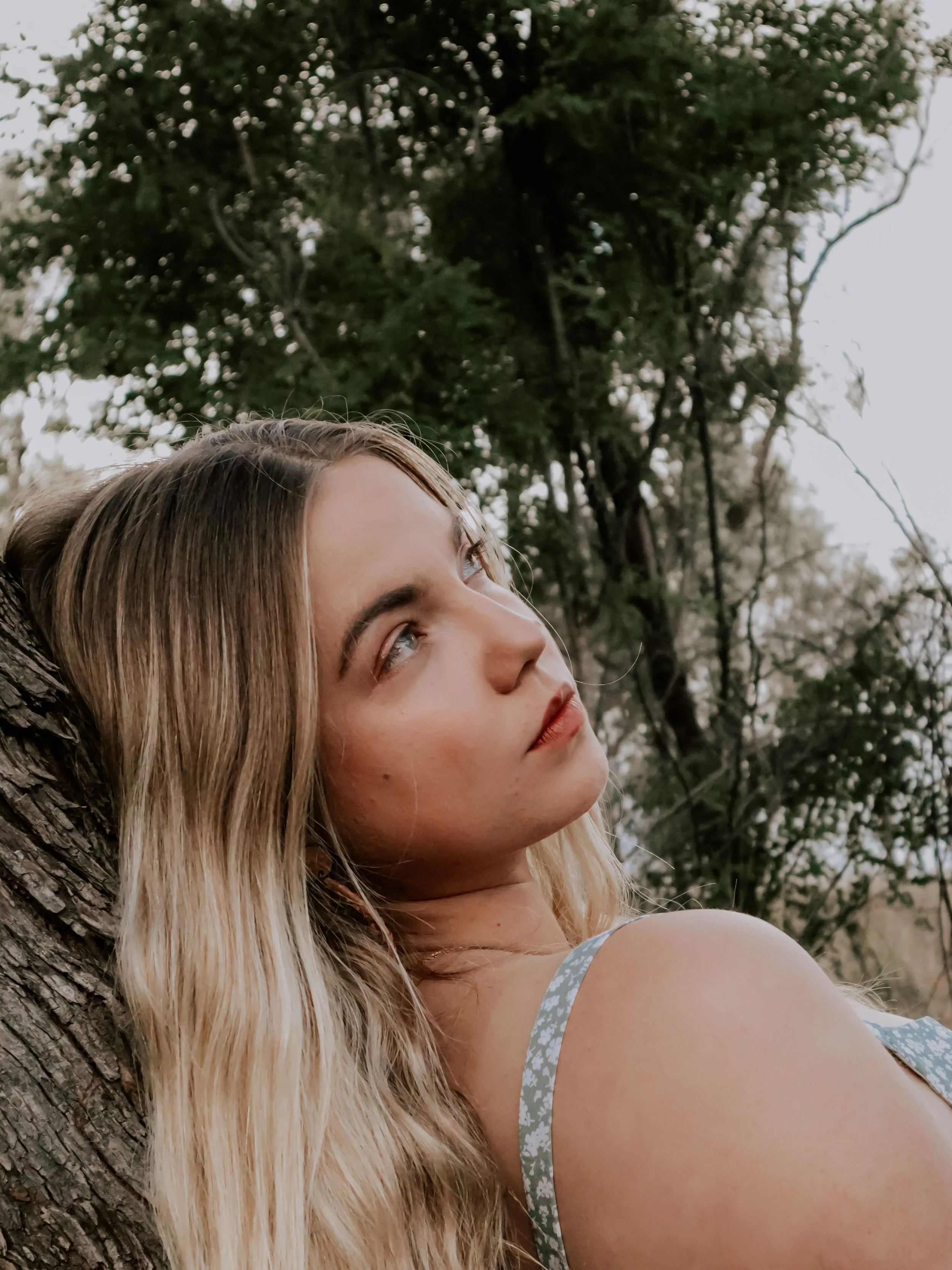 A young woman with blonde hair leaning against a tree, gazing thoughtfully into the distance with an outdoor forest background.