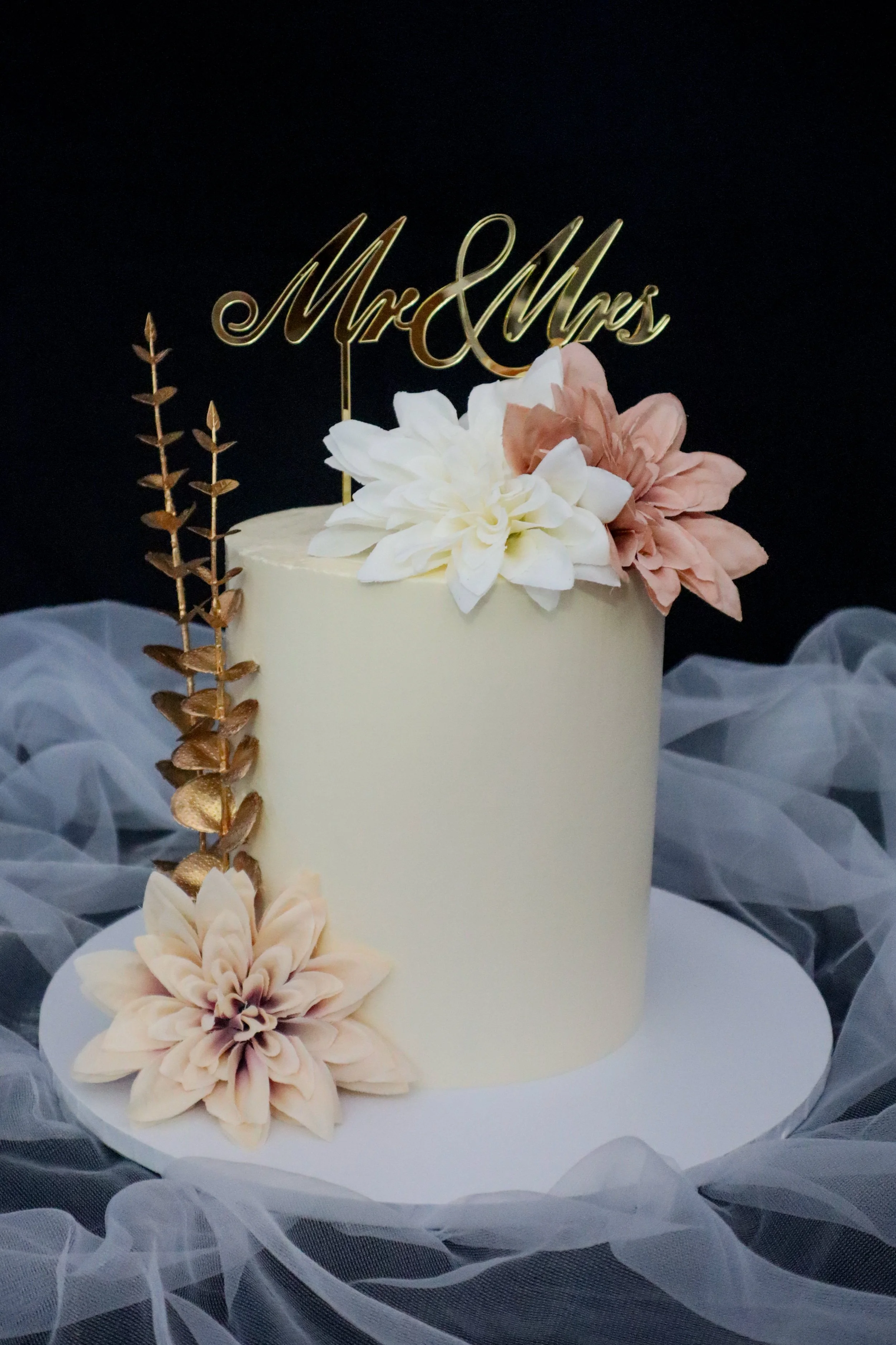Wedding cake decorated with flowers and a gold 'Mr & Mrs' topper, surrounded by white tulle fabric.