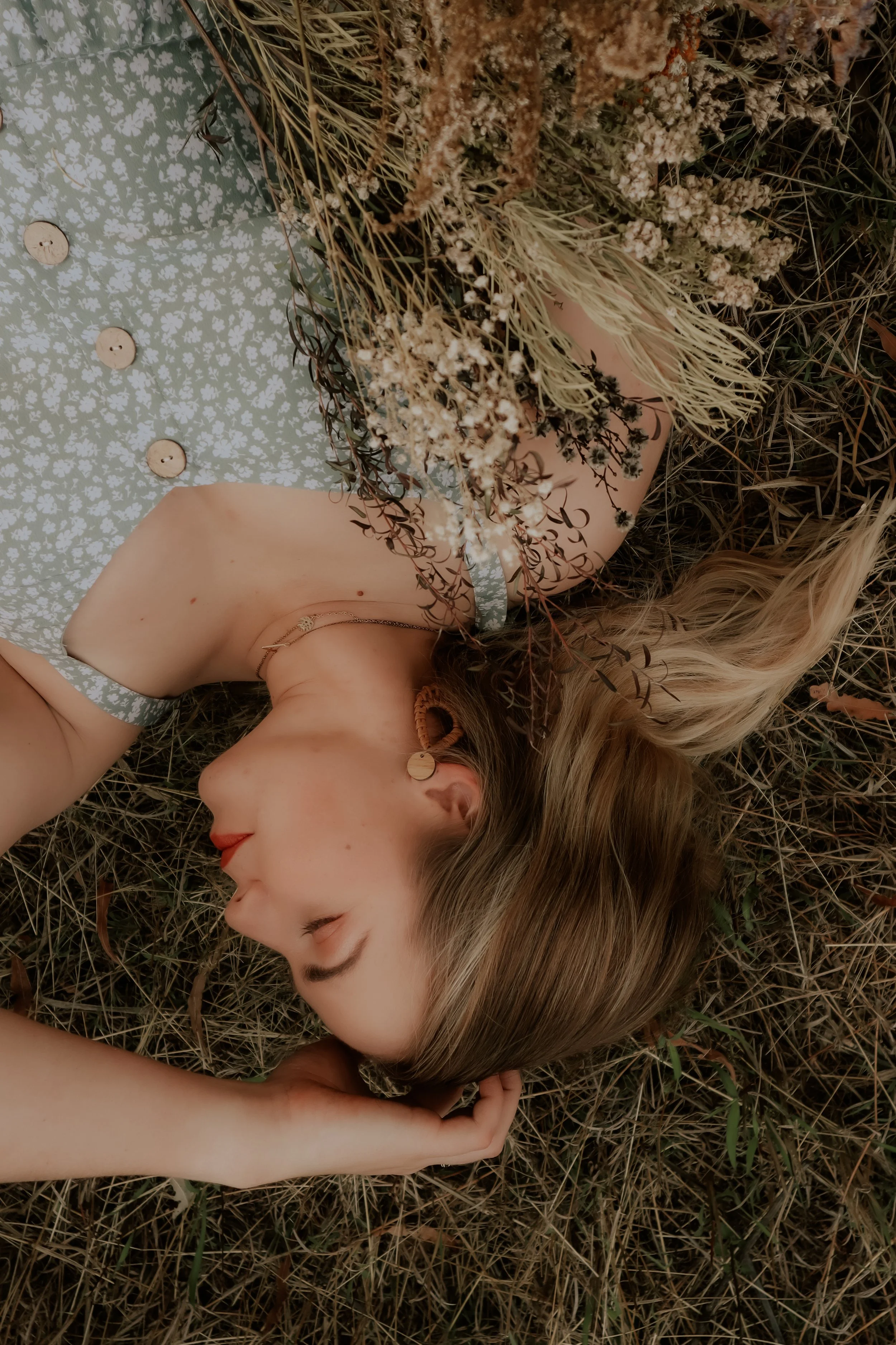 A woman with long blonde hair lying on the ground outdoors, surrounded by dry grass and small plants, with a bouquet of dry flowers covering part of her face.
