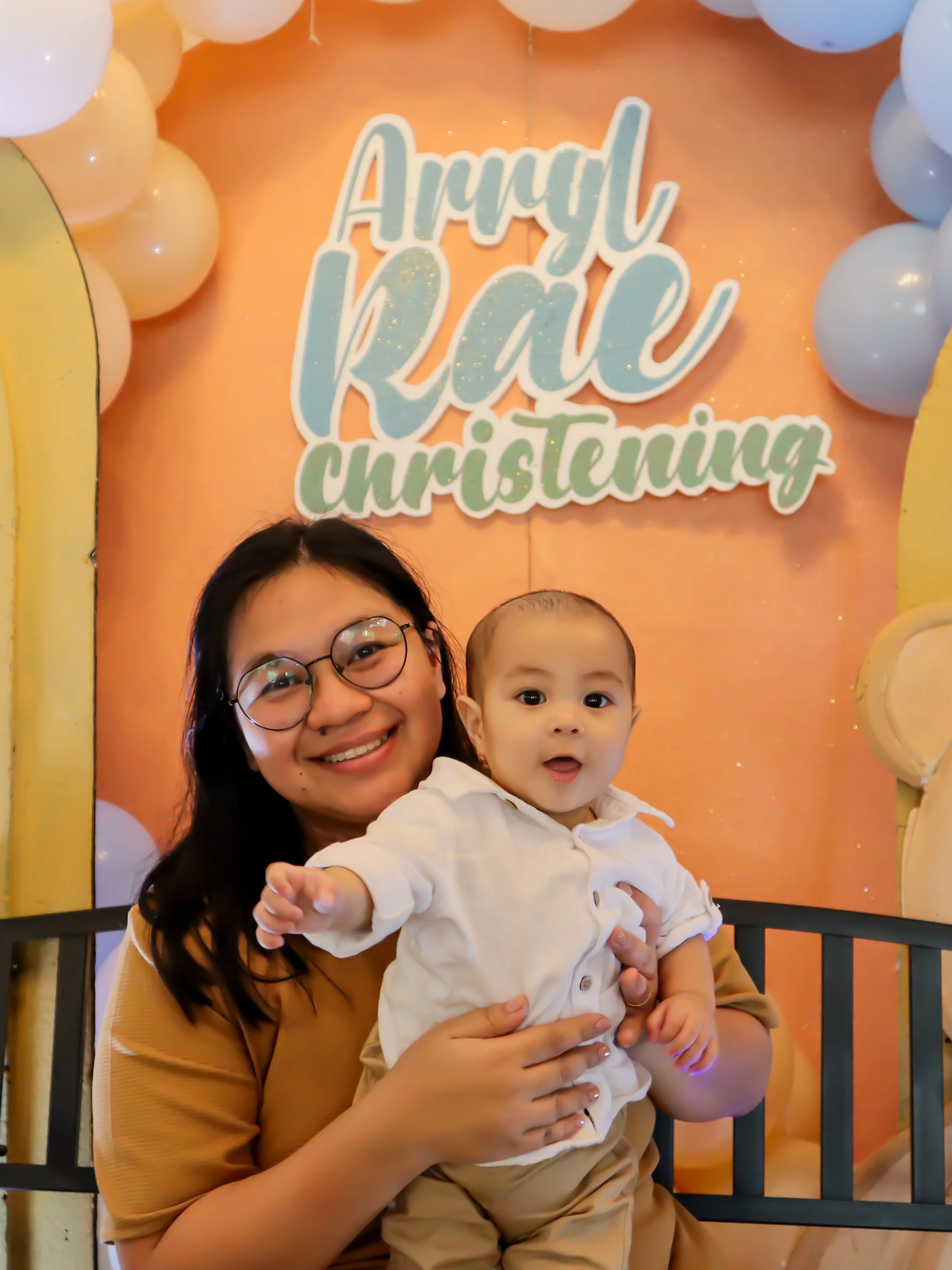 A woman wearing glasses holding a young boy at an anniversary and christening celebration. Behind them is a decorated backdrop with balloons and a sign that says 'Annual RAE Christening.'