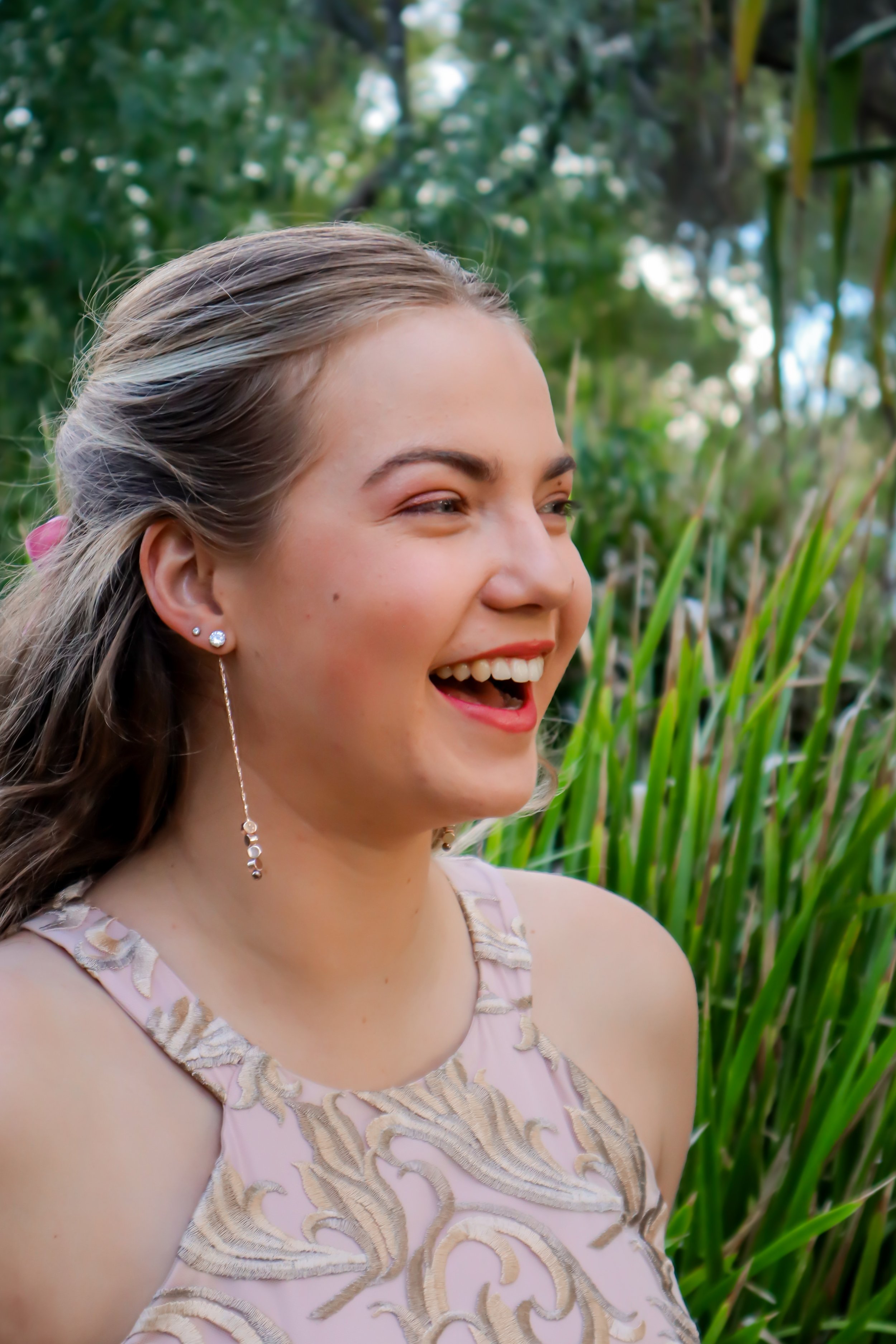 A young woman with blonde hair smiling outdoors among green plants, wearing a pink dress with gold embroidery and dangling earrings.