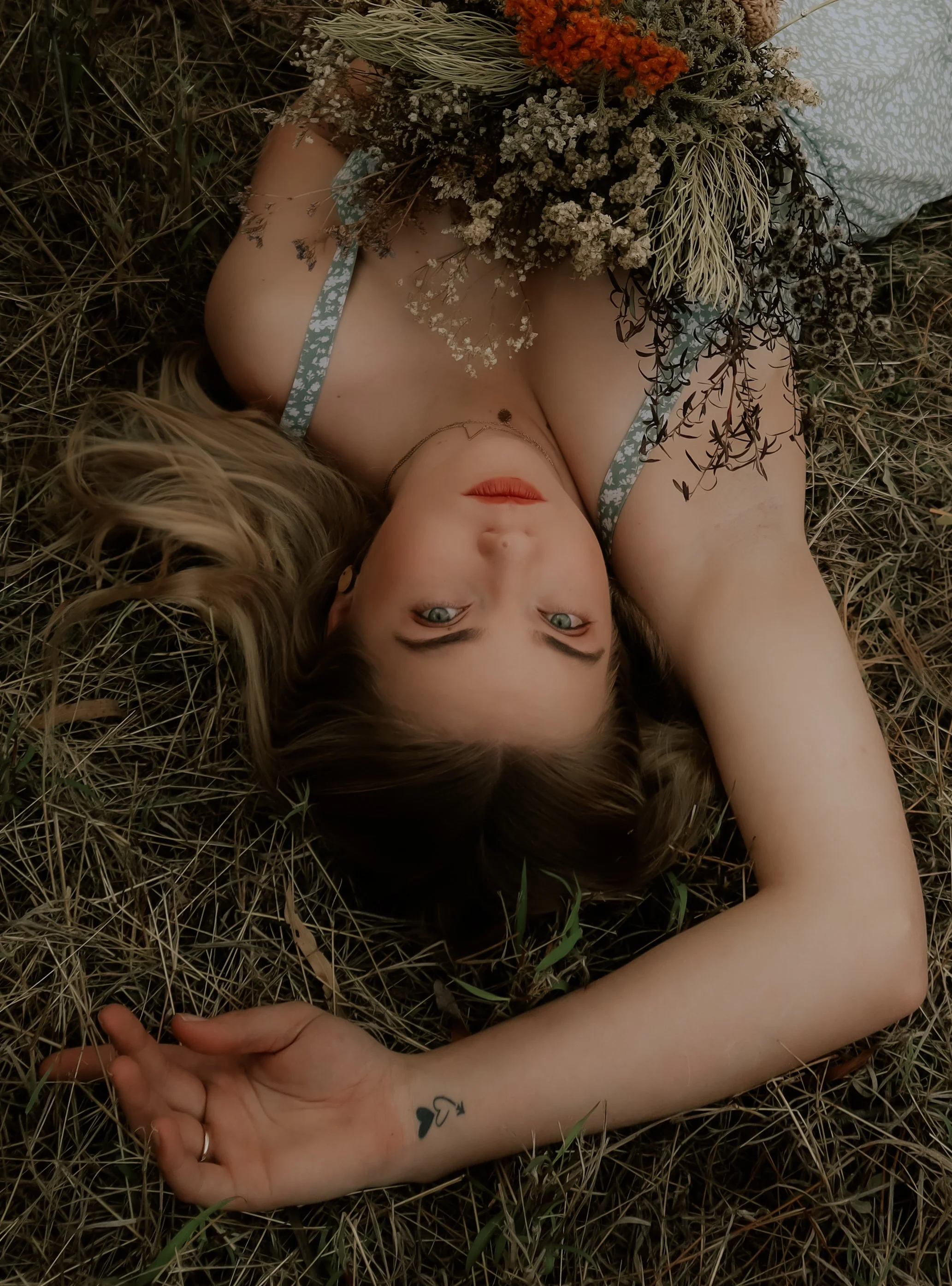 Young woman lying on the ground with a bouquet of dried flowers covering her chest, with her head tilted back and her arm extended, revealing a small tattoo on her wrist.