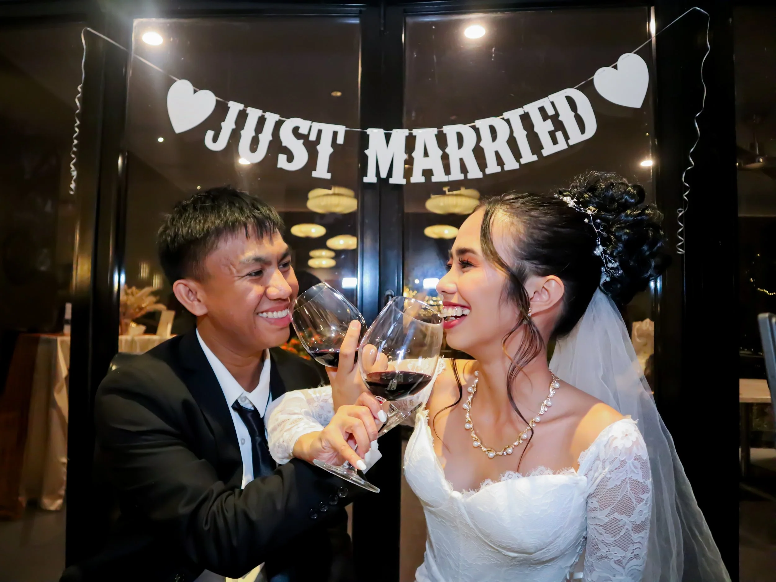 A newly married couple cheers with glasses of red wine in front of a window with a "Just Married" banner, celebrating their wedding night.