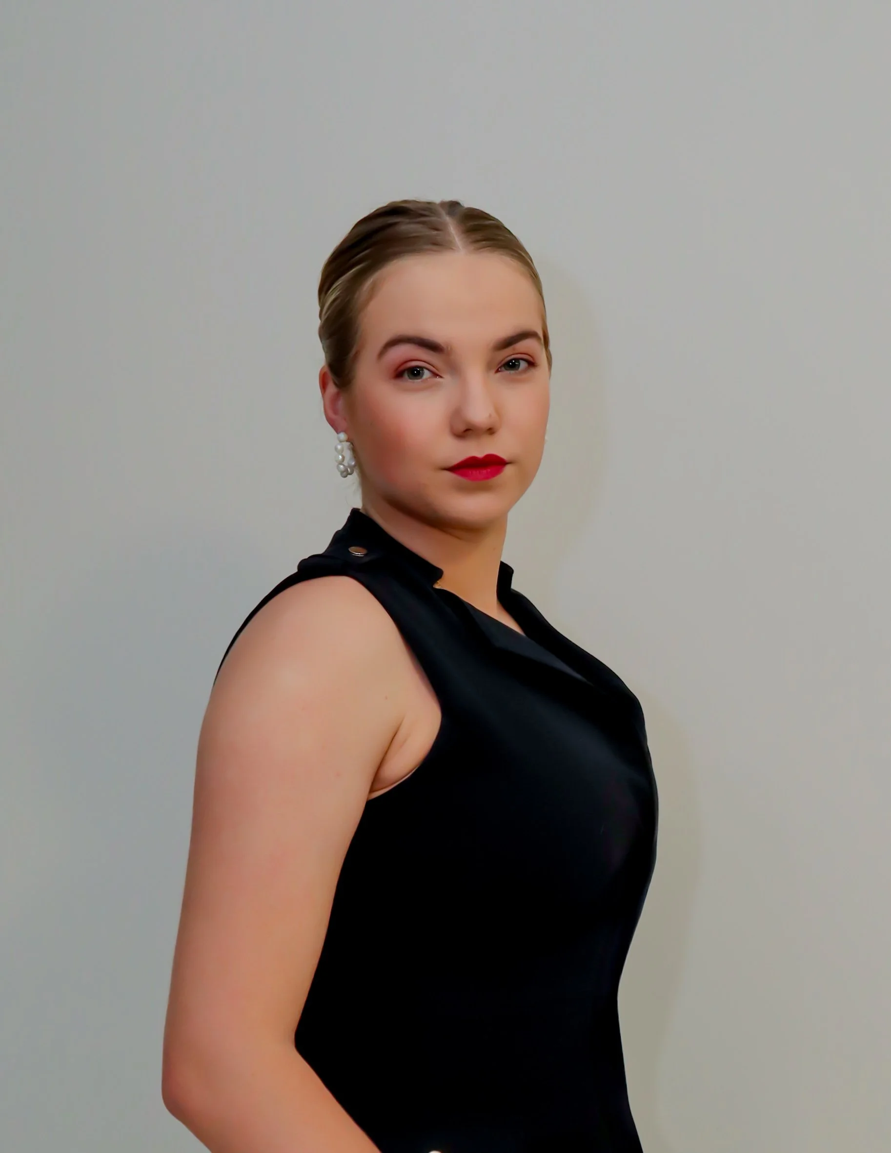 A young woman with blonde hair pulled back, wearing red lipstick, pearl earrings, and a black sleeveless dress with a high collar, standing against a plain white wall.