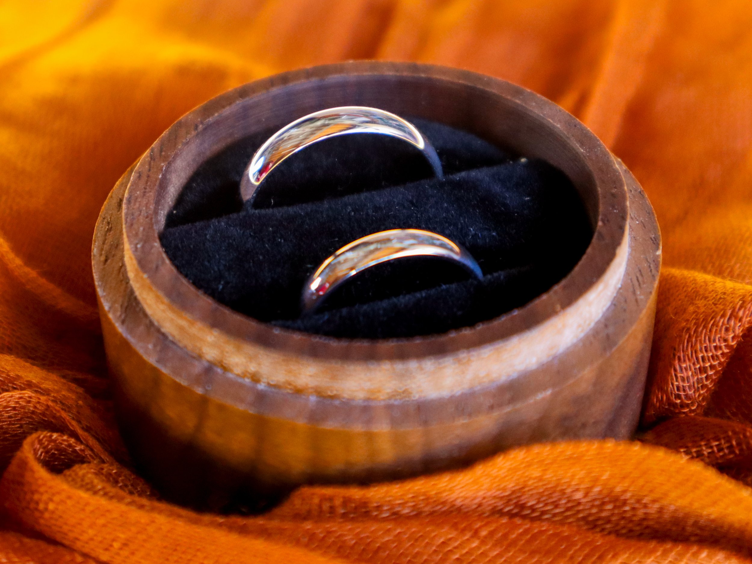 A wooden ring box with two silver wedding bands inside, resting on orange fabric.