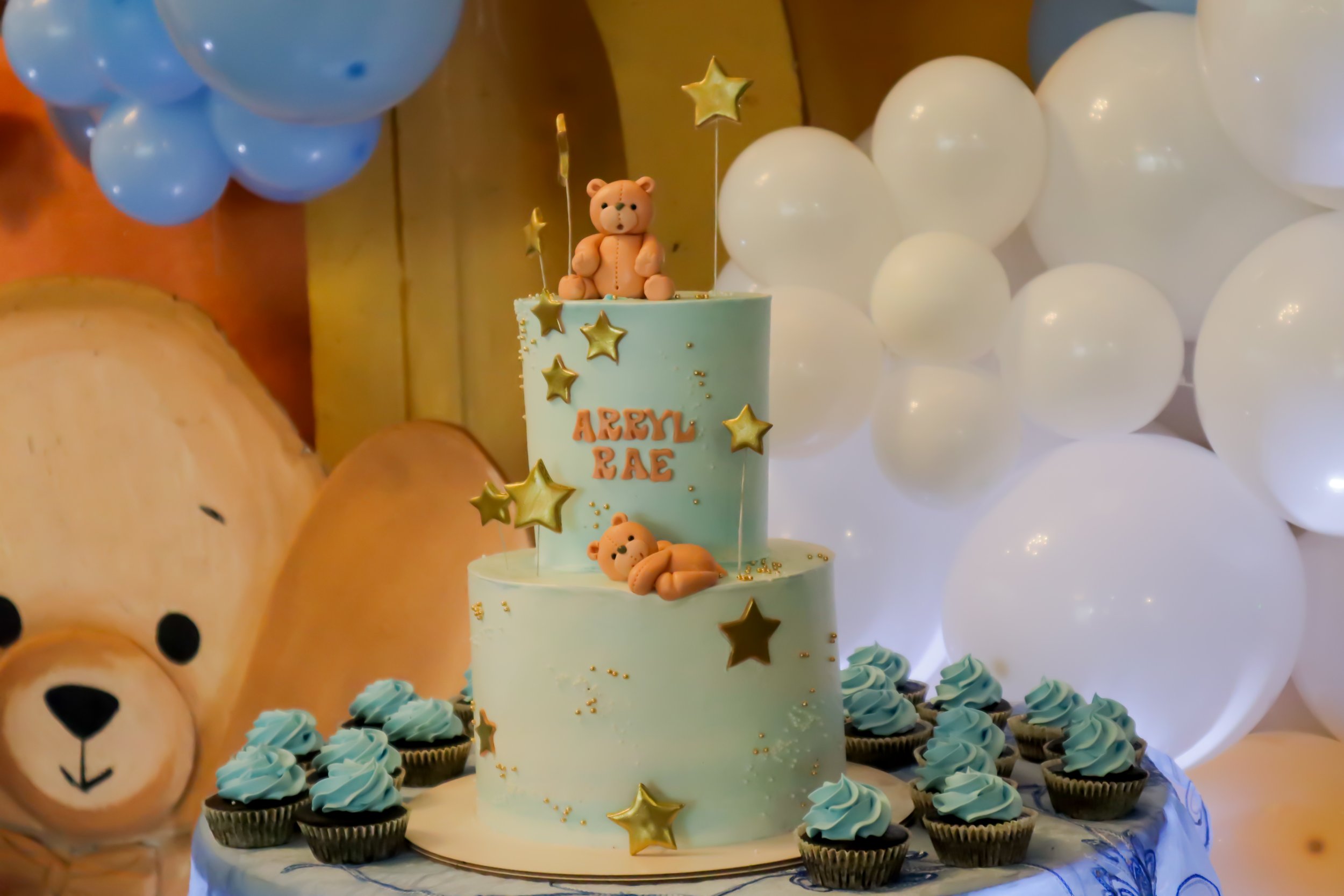 A two-tiered light blue birthday cake decorated with golden star toppers, with teddy bear toppers on top and nestled on the side, surrounded by blue frosted cupcakes. In the background, white balloons and a large teddy bear's face backdrop are visibl