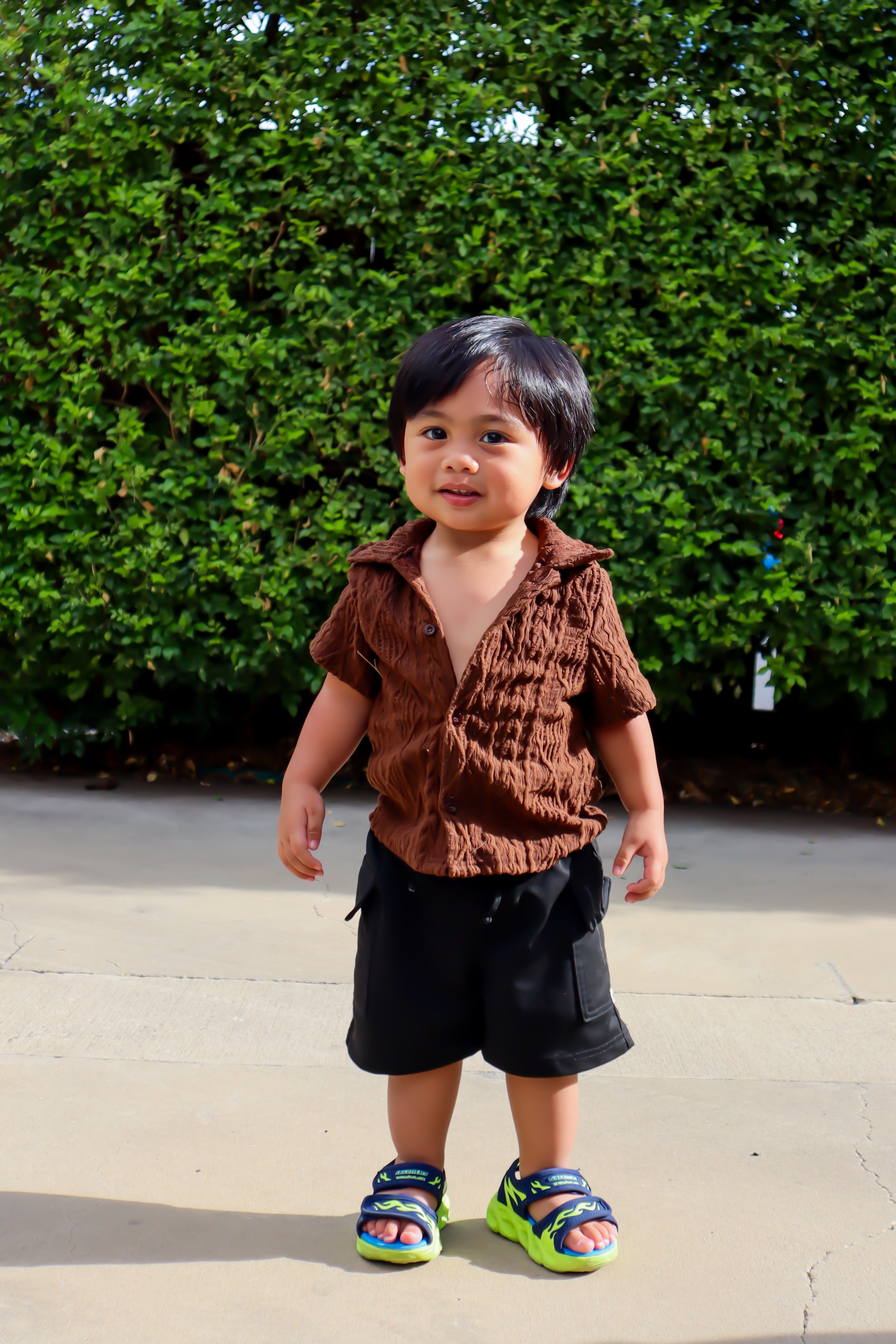 A young child with dark hair and a light skin tone standing outdoors in front of a green hedge, wearing a brown textured short-sleeve shirt, black shorts, and bright green and blue sandals.