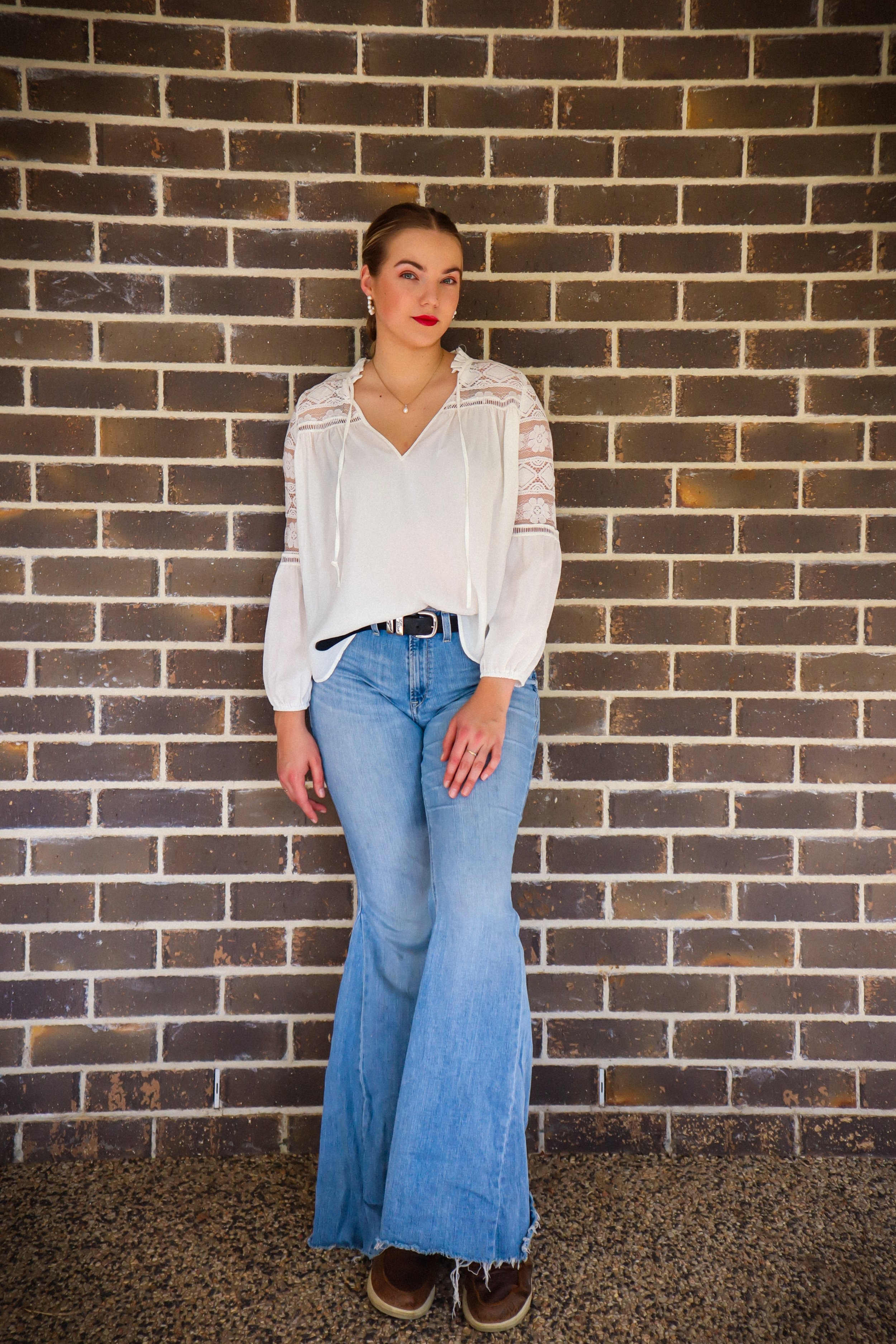 A young woman with fair skin, red lipstick, and brown hair in a braid, standing against a brick wall, wearing a white lace blouse, blue jeans with frayed hems, and brown shoes.